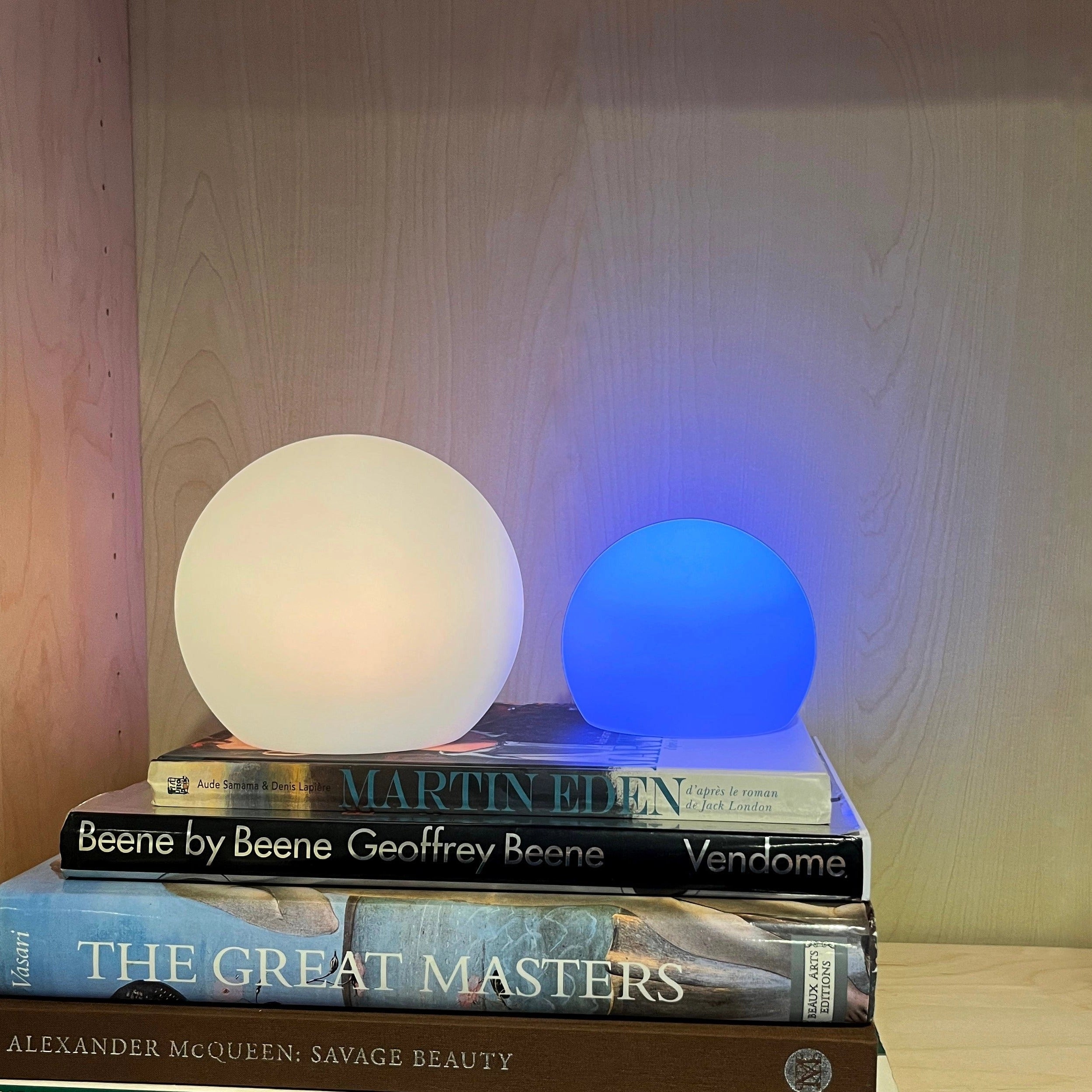 Modern minimalist LED globe lamp with rechargeable cordless design, placed on a wooden rectangular bedside table with shelving against a hardwood wall.