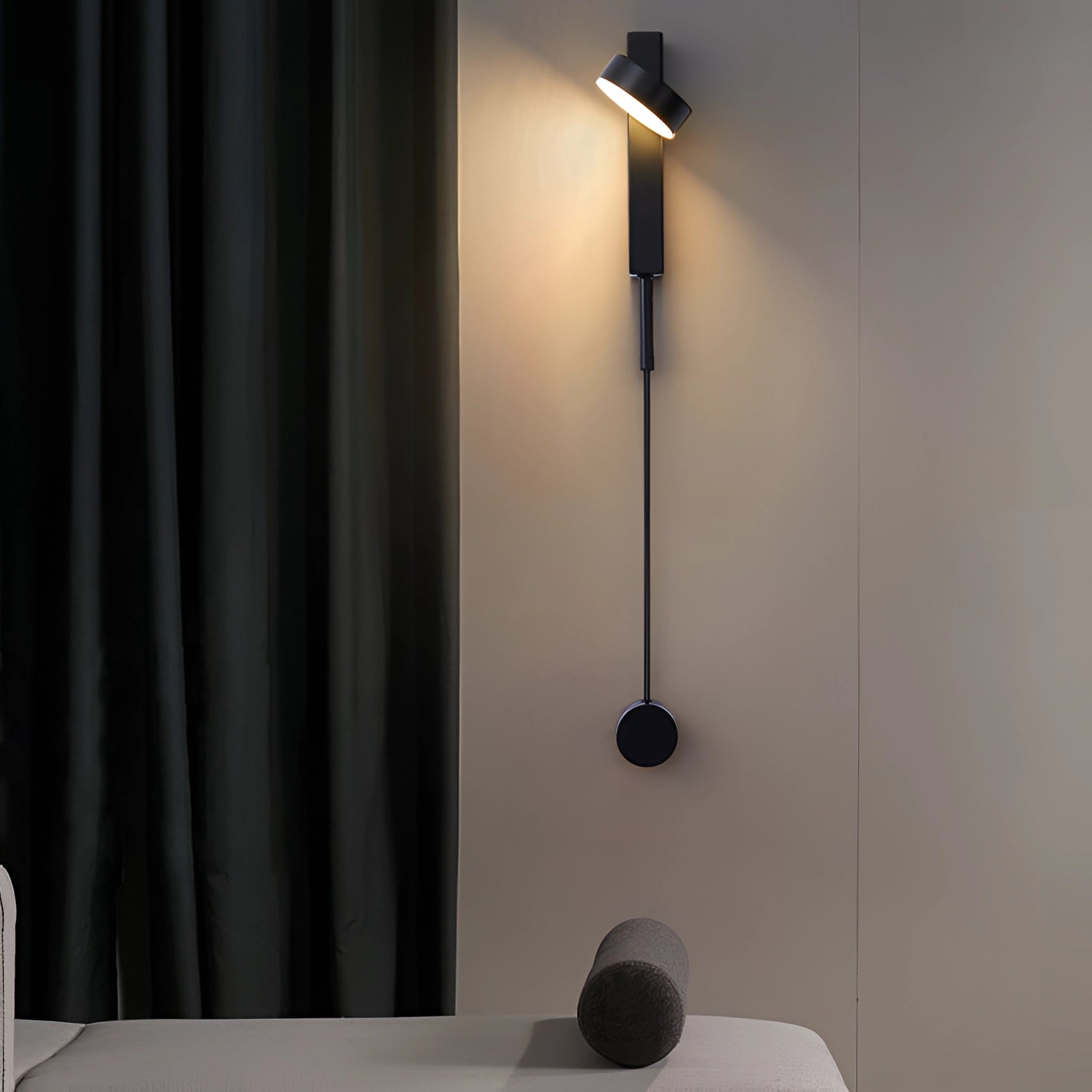 Modern minimalist LED wall sconce in black with an adjustable design, mounted on a wall in a warmly lit bedroom setting. The sconce emits a soft warm light (3000K) suitable for UK/Australia voltage (220 V-240 V), complementing the contemporary interior decor featuring wooden flooring, curtains, and a neutral color palette.