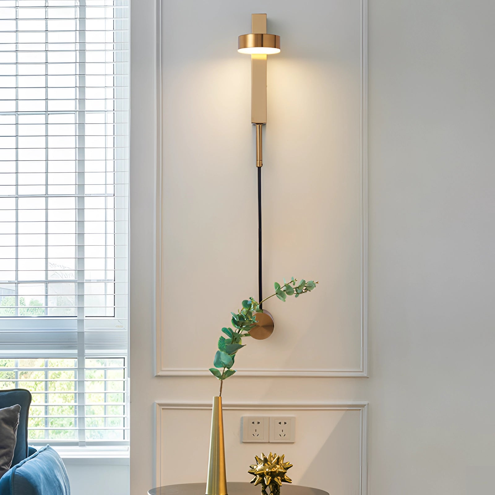 Modern minimalist LED wall sconce in gold, mounted on a wooden wall beside a window, providing warm 3000K light to a stylishly decorated bedroom with houseplants in flowerpots and contemporary interior design elements.