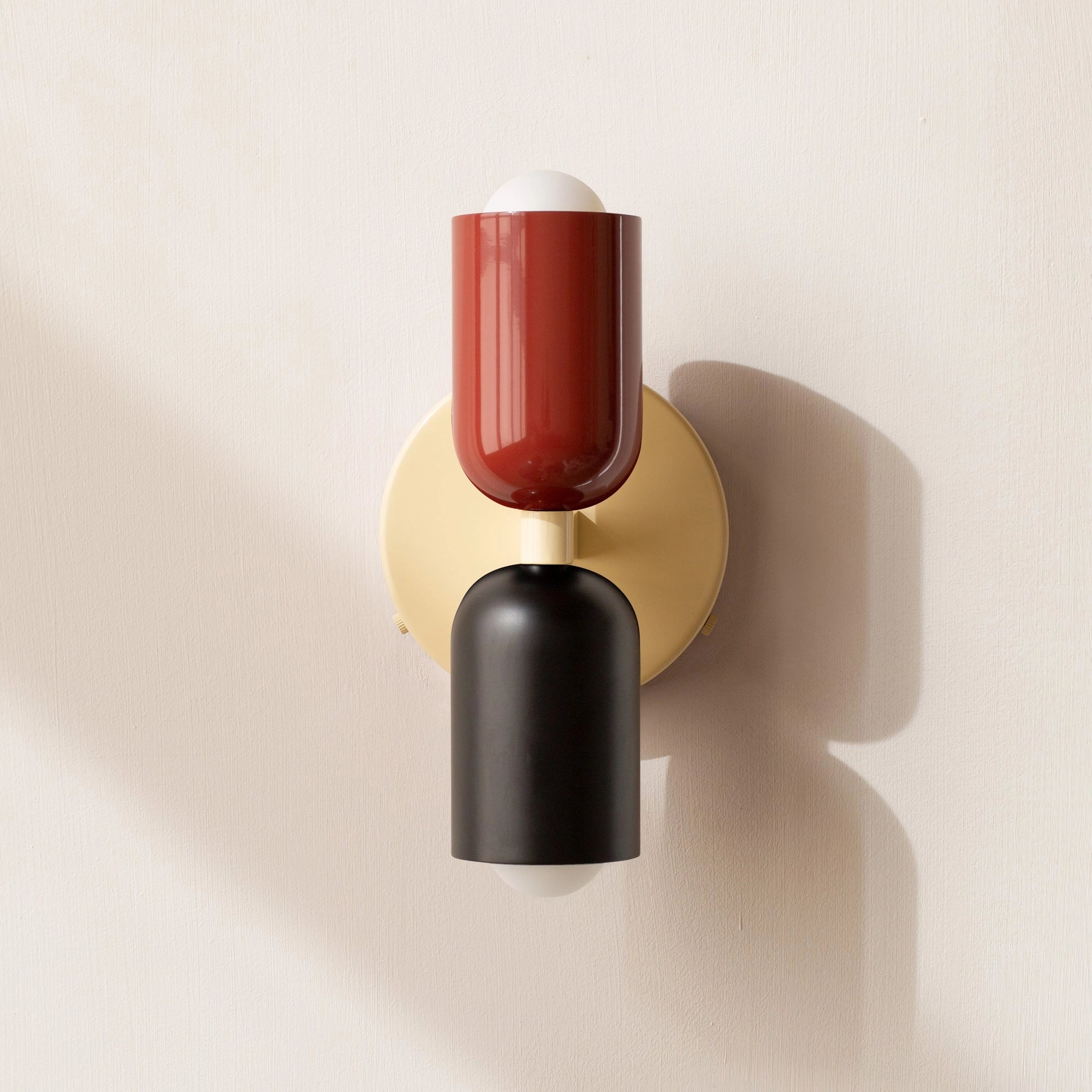 A modern minimalist LED wall sconce featuring a geometric dual head lamp design with a blend of red, black, and beige colors, highlighted by its sleek, cylindrical metal structure against a wooden background.
