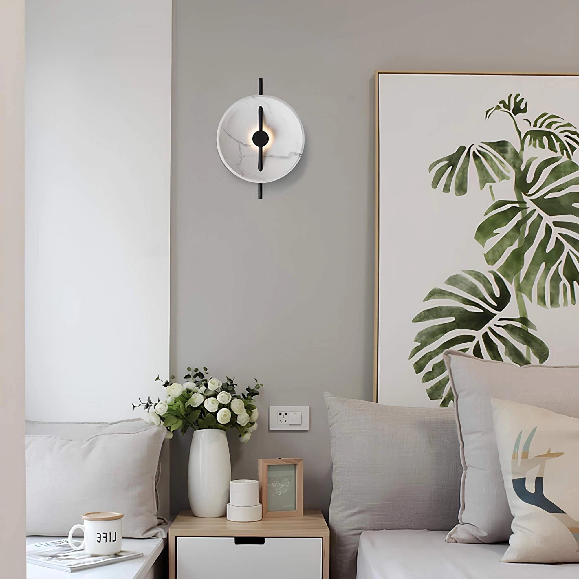Modern minimalist silver round wall clock hanging on a white wall in a stylishly decorated interior featuring a grey couch, a plant, and a picture frame.