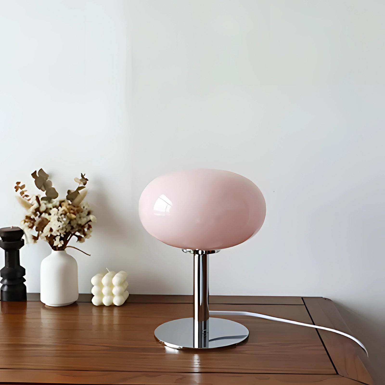Modern minimalist white globe table lamp with a chrome base on a wooden table, featuring a pink accent, surrounded by contemporary interior design elements, including a vase with a plant.