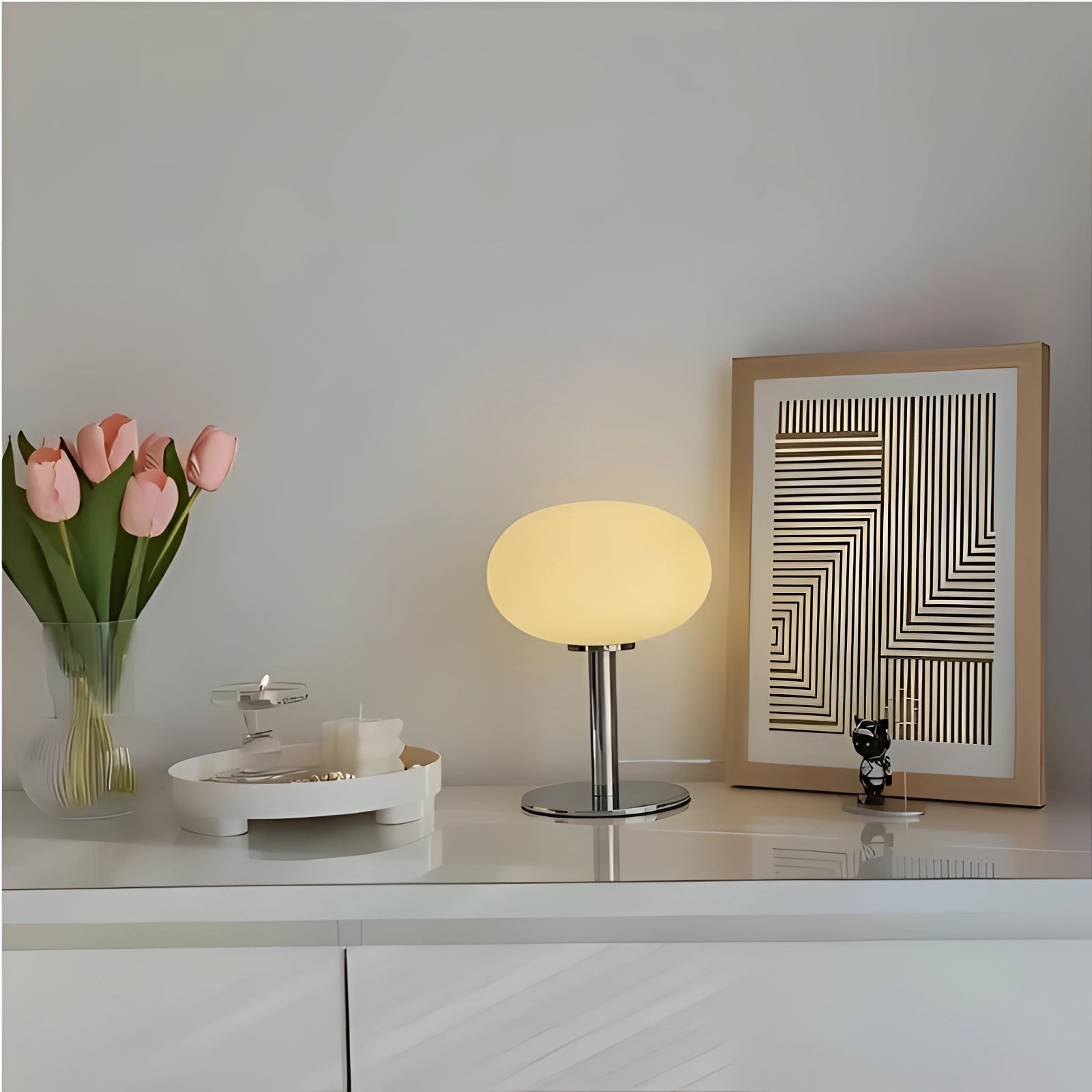 A modern minimalist white globe table lamp with a chrome base, set against a wooden background, featuring a yellow tint. The lamp is part of an elegant, contemporary interior design display, complemented by a nearby plant and decorative elements.