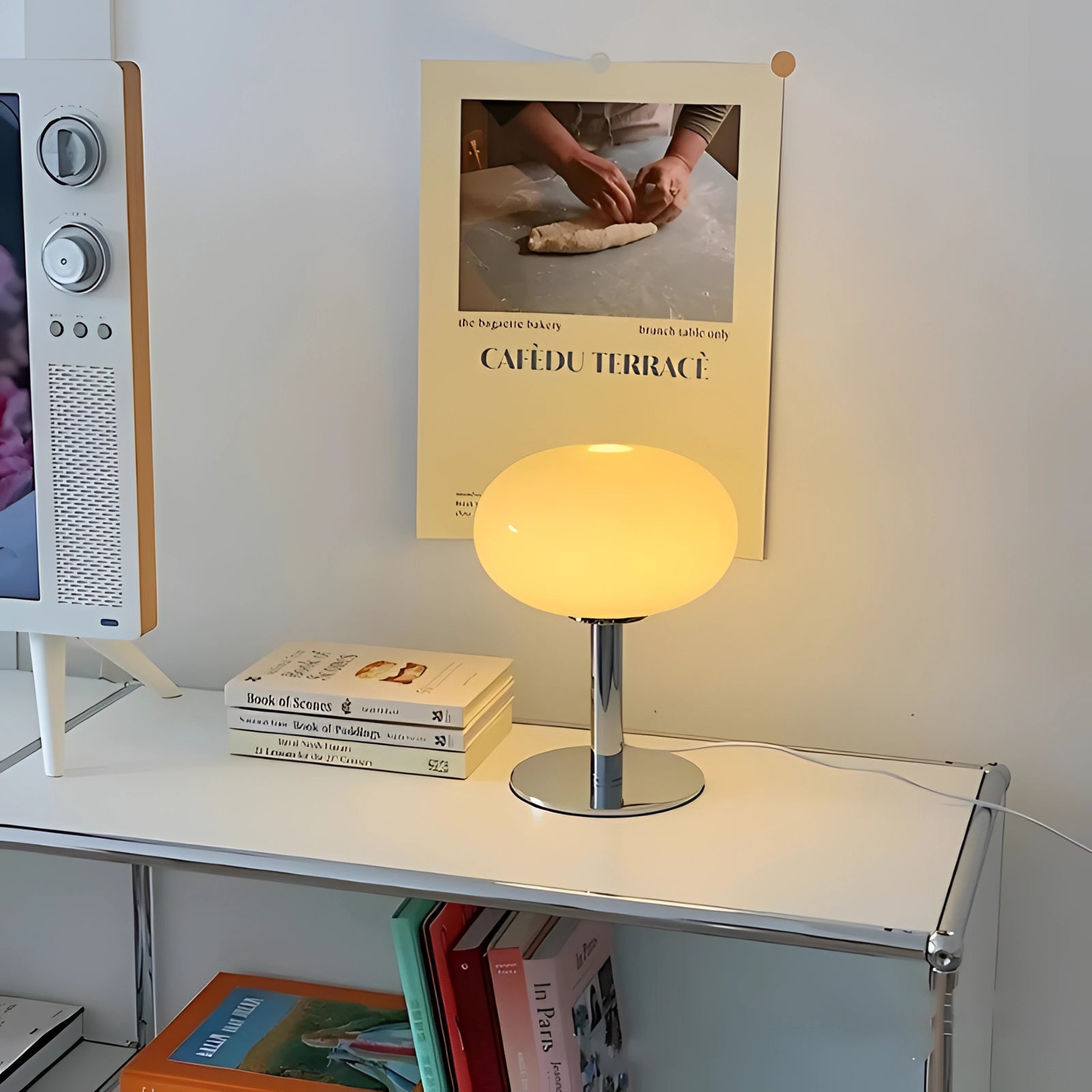 Modern minimalist white globe table lamp with a chrome base placed on a wooden desk next to a bookcase filled with books and decorative items.