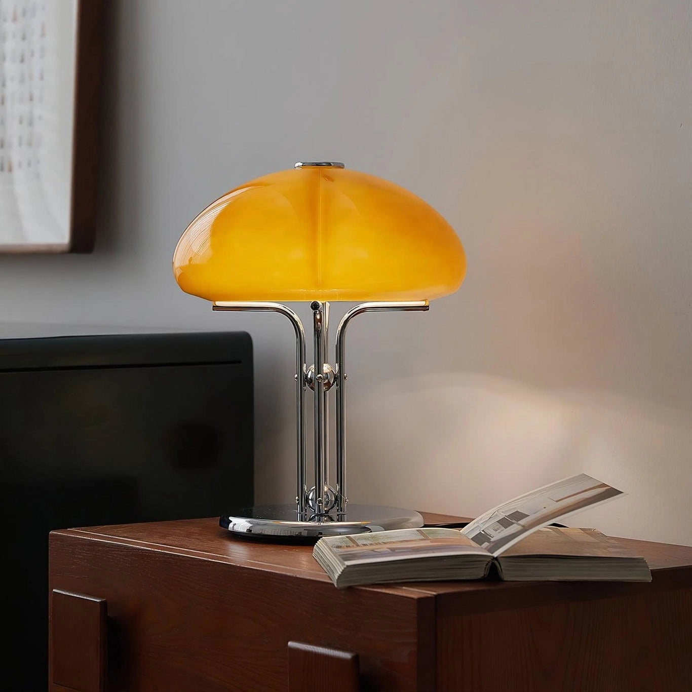 Modern orange globe table lamp with chrome base emitting warm light, placed on a wooden table next to a plant on a shelf.