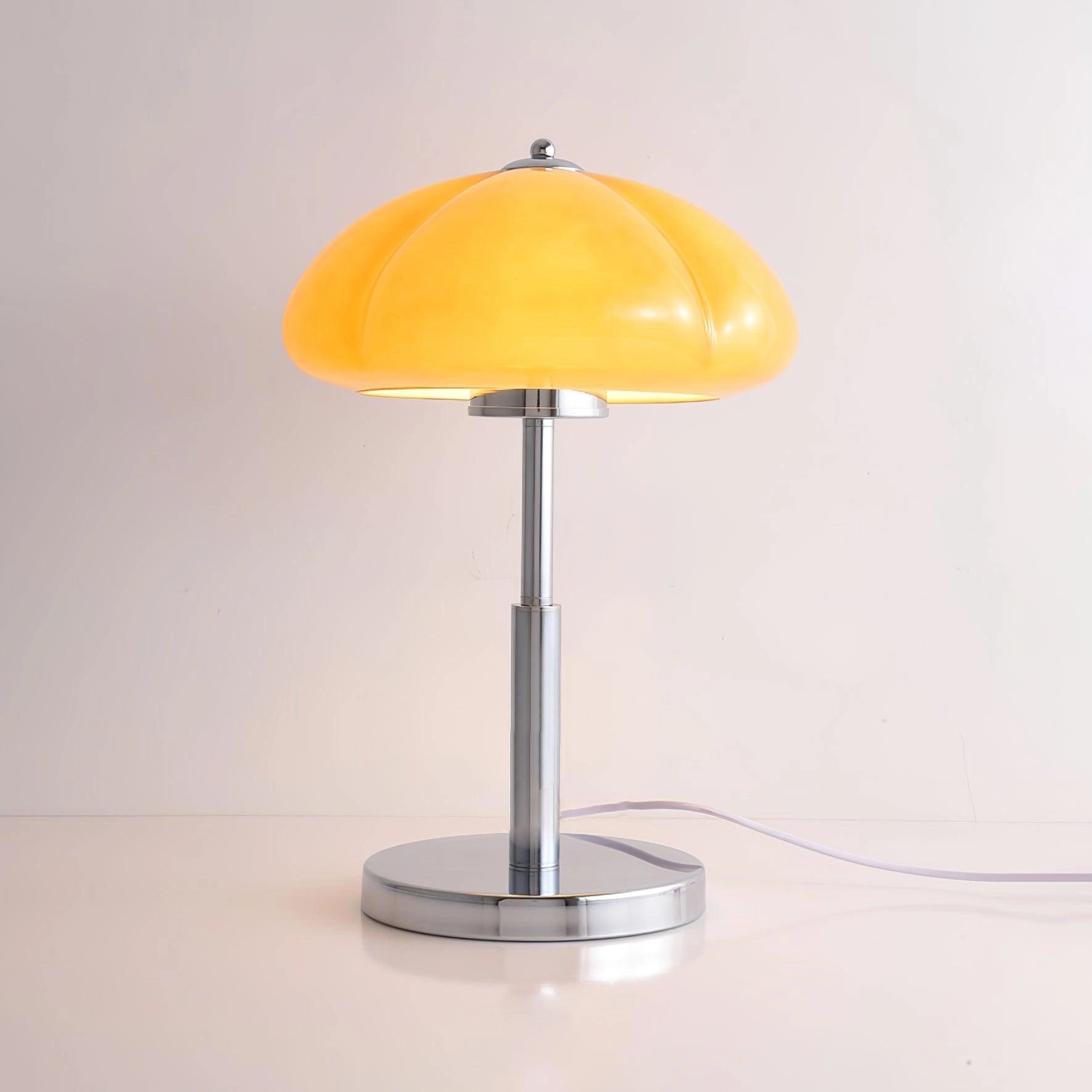 Modern orange globe table lamp with chrome base on a wooden table, emitting warm light, decorative accent with plant in the background.