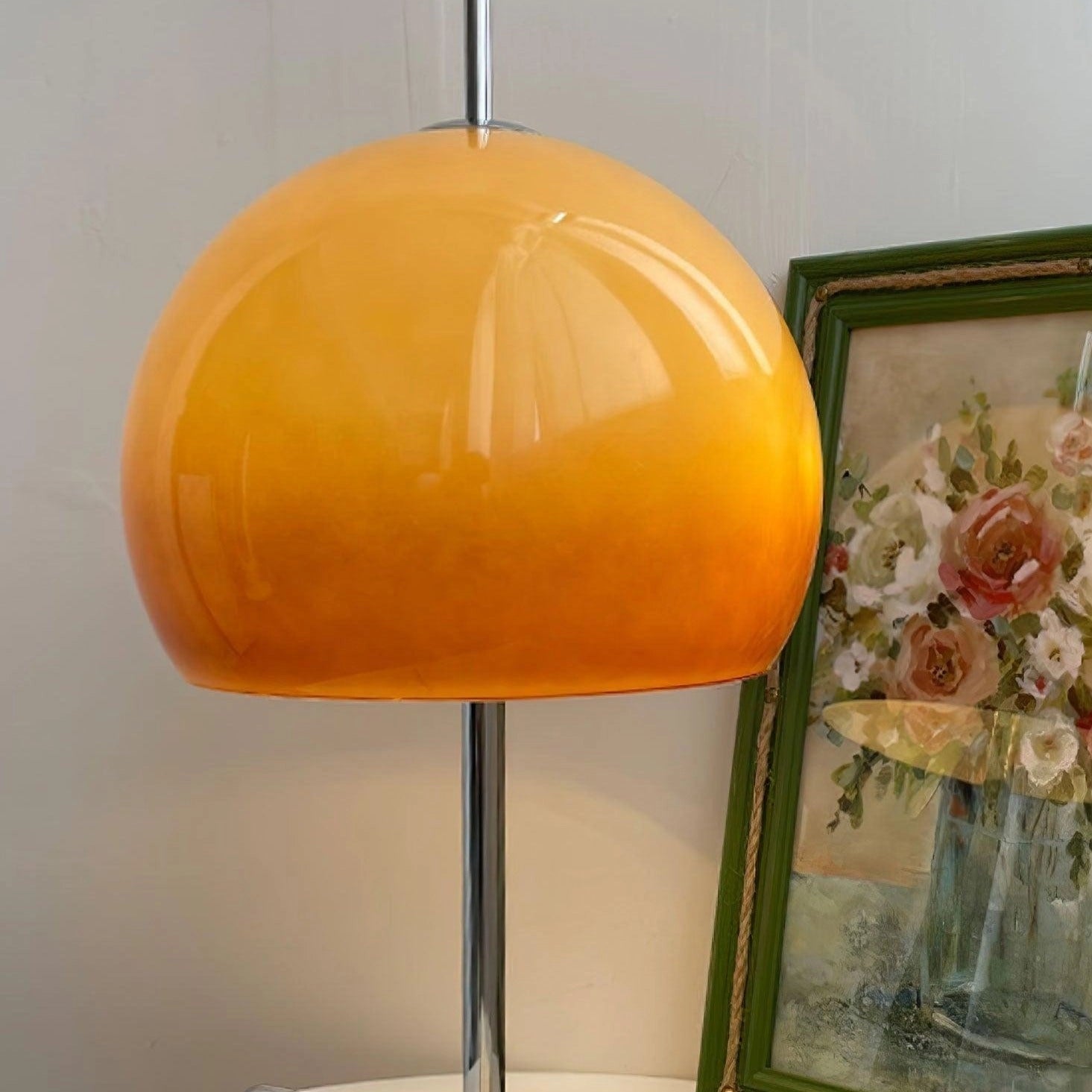 Modern orange globe table lamp with chrome base emitting warm light, placed on a wooden table next to a chair, picture frame, and decorative flowers.