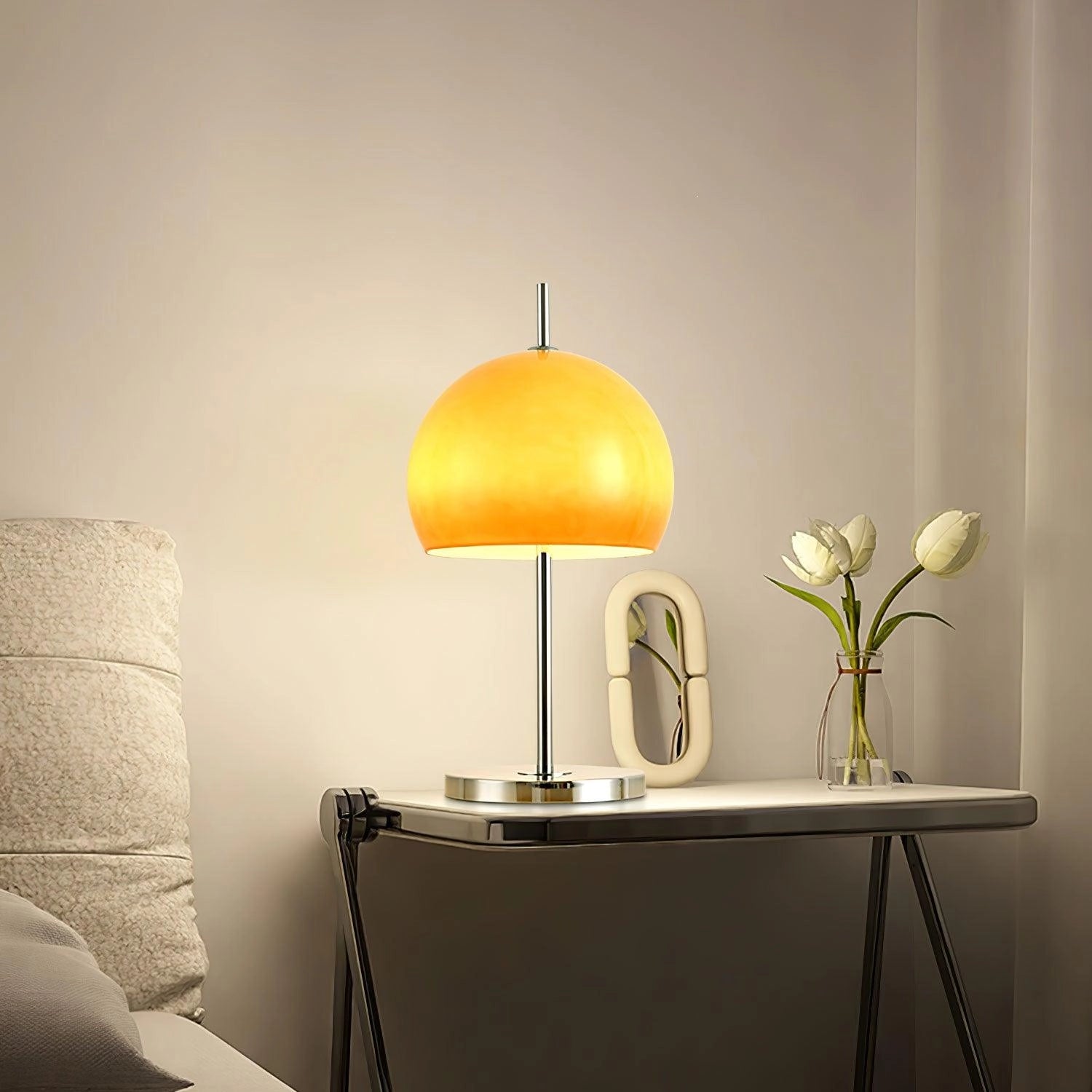 Modern orange globe table lamp with chrome base, emitting warm light, placed on a wooden table in a stylish interior setting.
