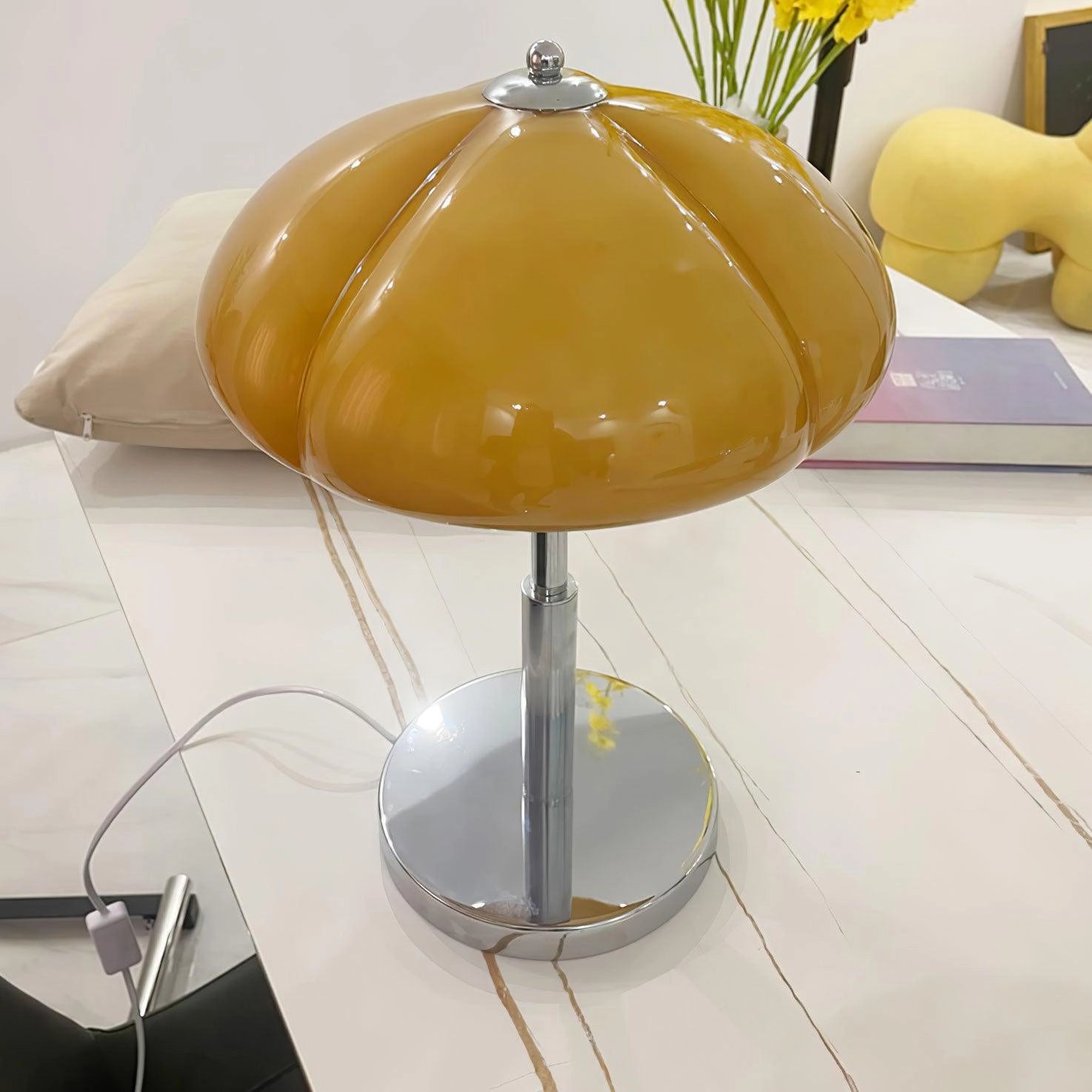 Modern orange globe table lamp with chrome base emitting warm light, placed on a wooden table next to a potted plant.