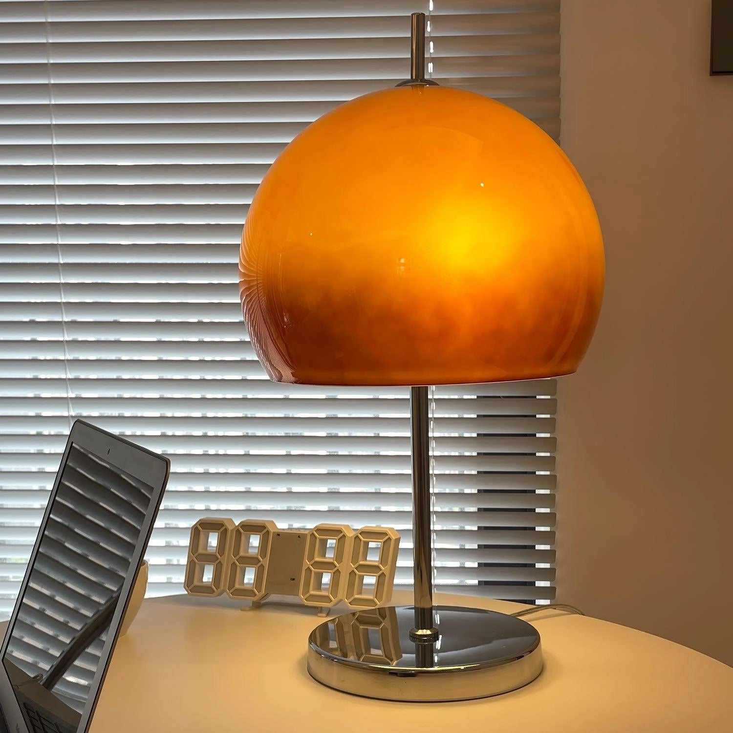Modern orange globe table lamp with chrome base, emitting warm light, placed on a wooden table in an interior setting.