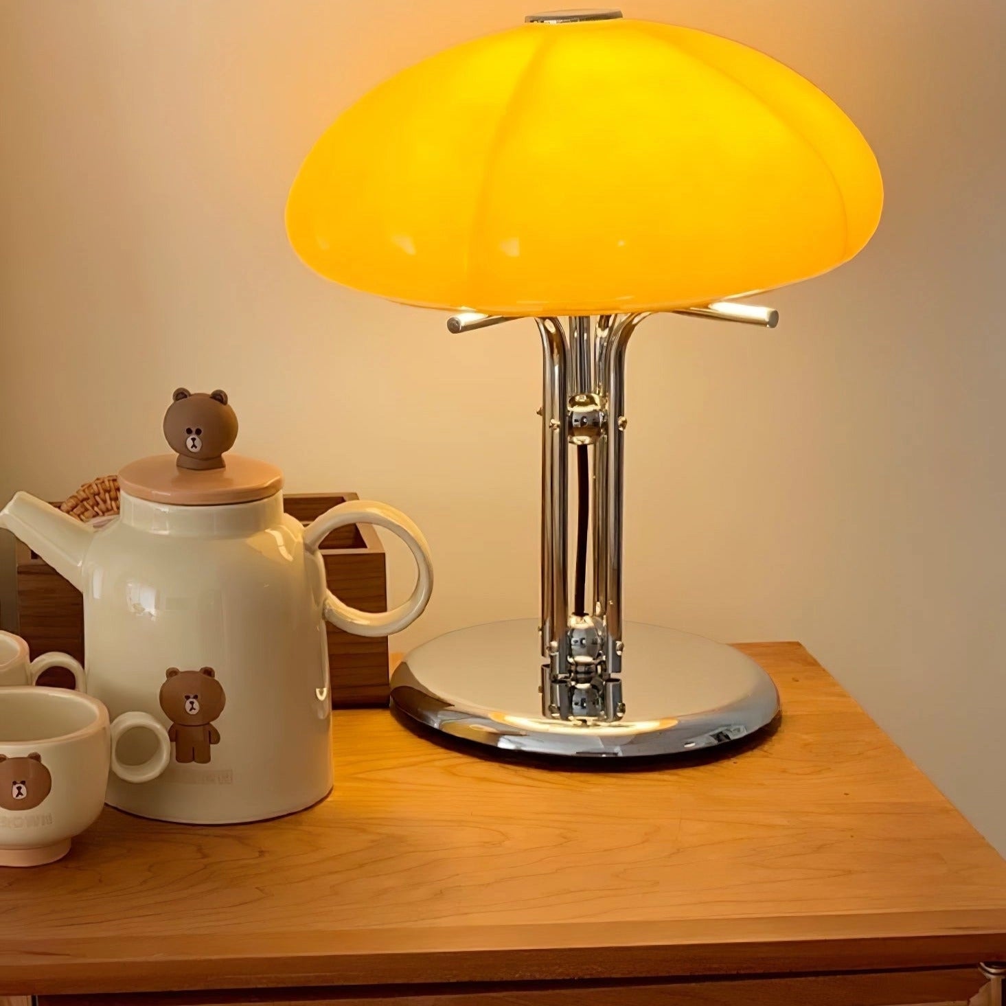 Modern orange globe table lamp with chrome base, emitting warm light, placed on a wooden table with various tableware and drinkware items.
