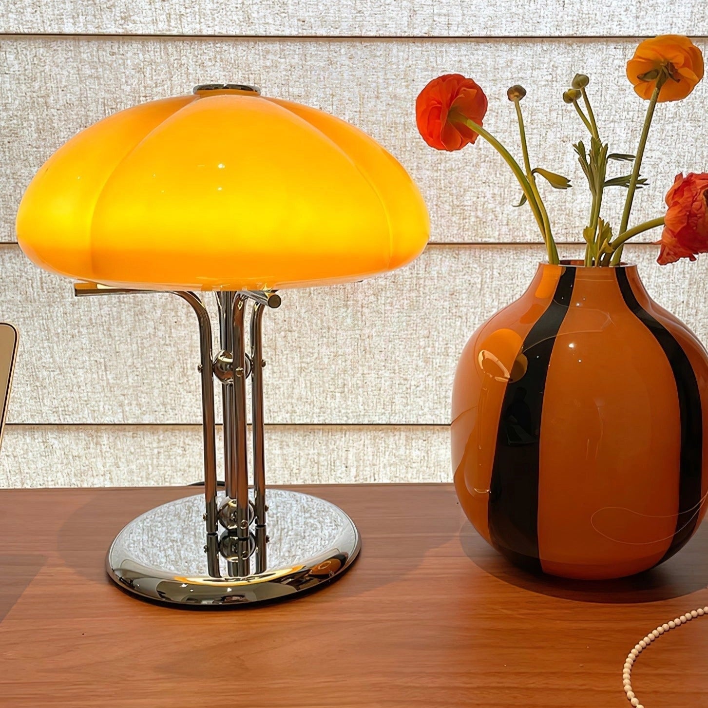 Modern orange globe table lamp with chrome base emitting warm light, placed on a table next to a vase with flowers.
