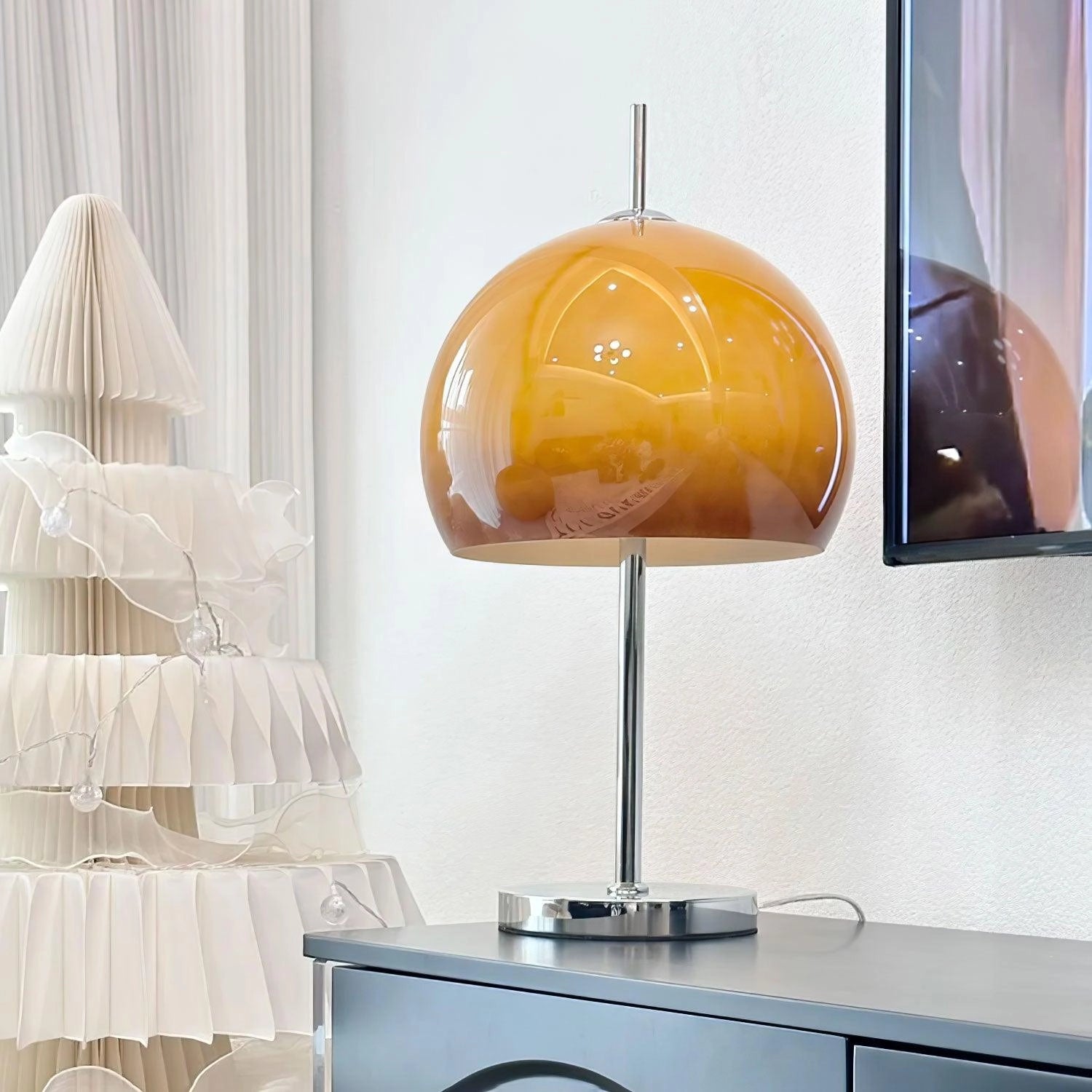 Modern orange globe table lamp with chrome base emitting warm light, placed on a table next to a television and shelving units in a contemporary styled room.