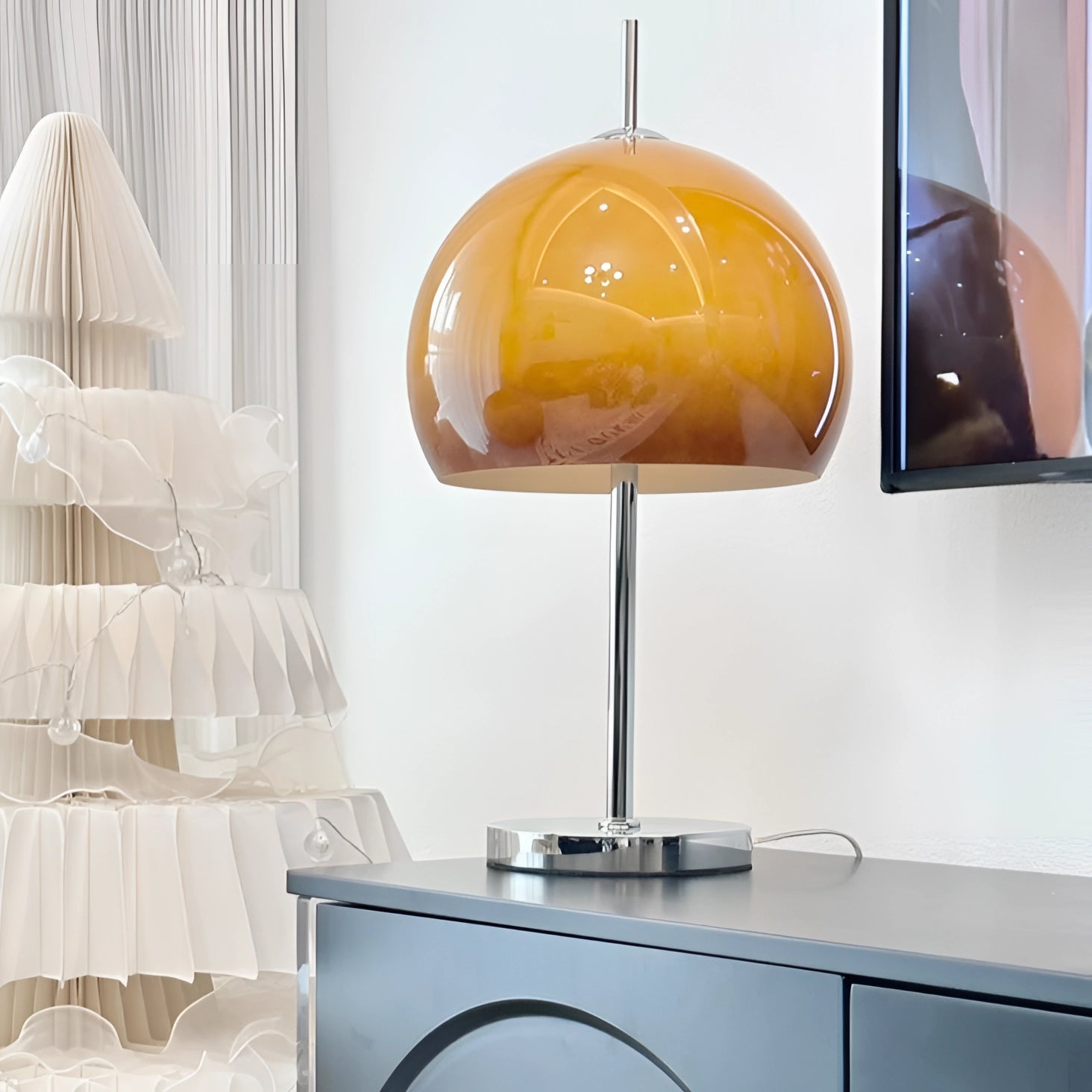 Modern orange globe table lamp with chrome base emitting warm light, placed beside a television on a shelving unit in a stylish interior design setting.