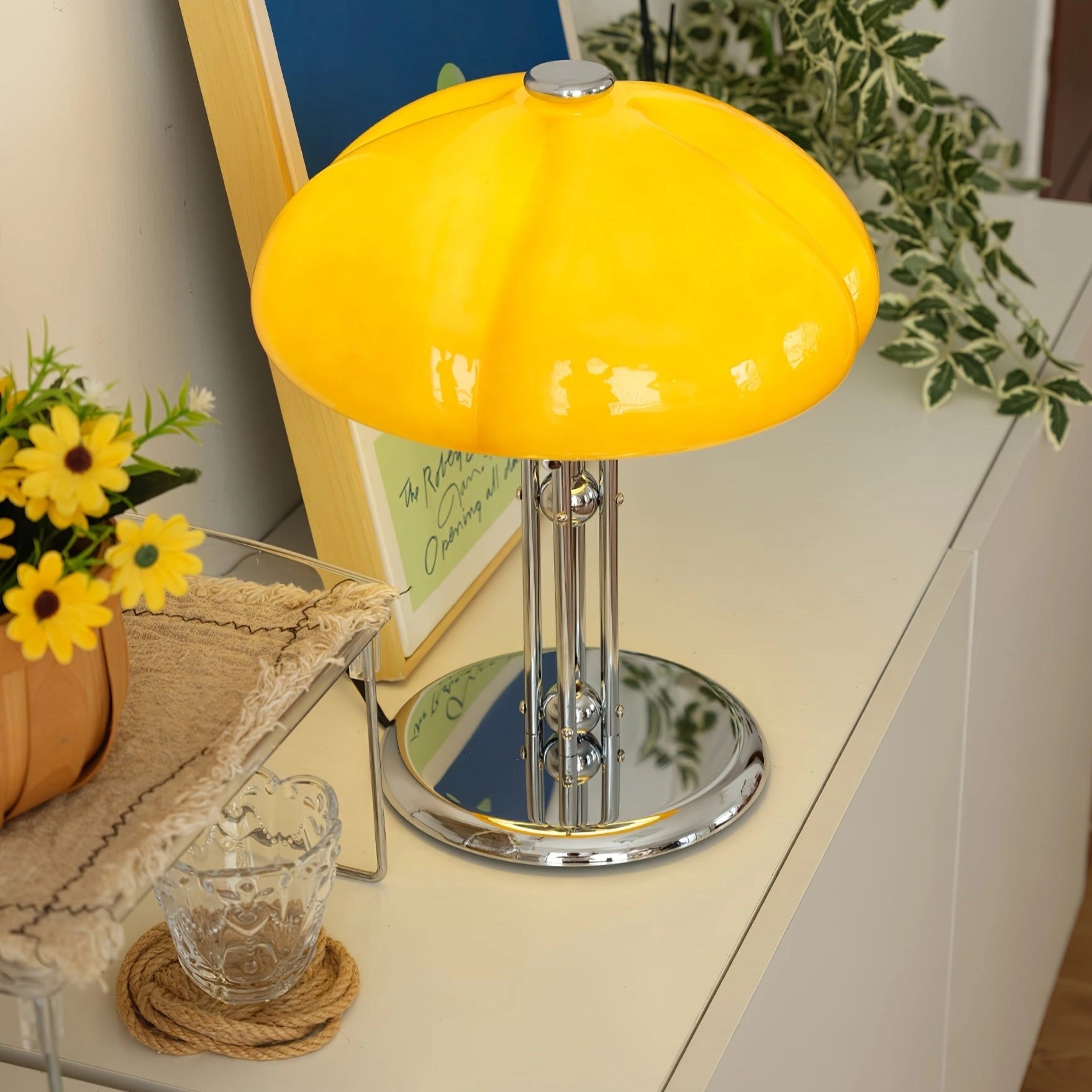 Modern orange globe table lamp with chrome base emitting warm light, placed on a table with decorative elements like a plant, tableware, and a flower, creating a cozy interior setting.