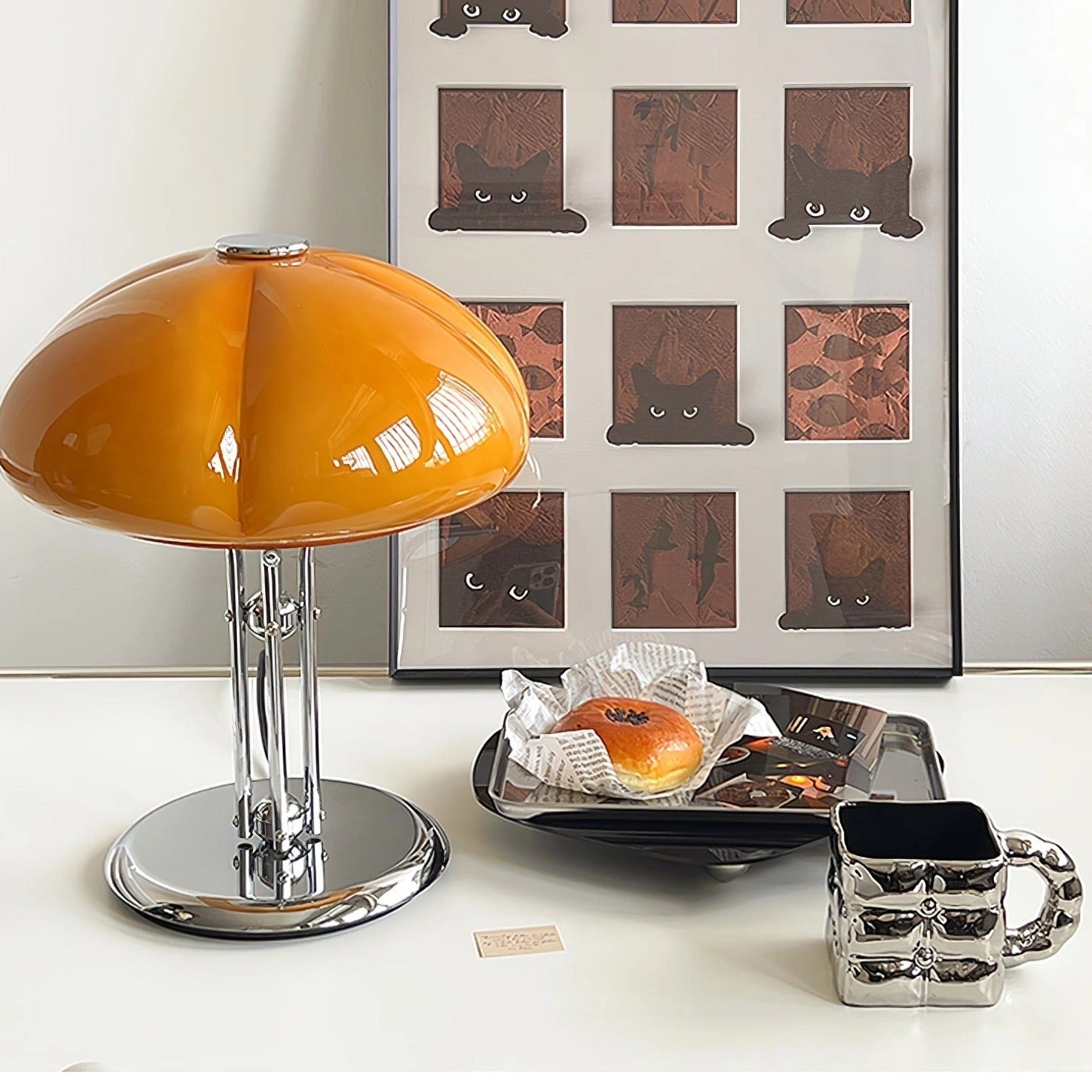Modern orange globe table lamp with chrome base, emitting warm light, placed on a table next to decorative plants, showcasing interior design elements.
