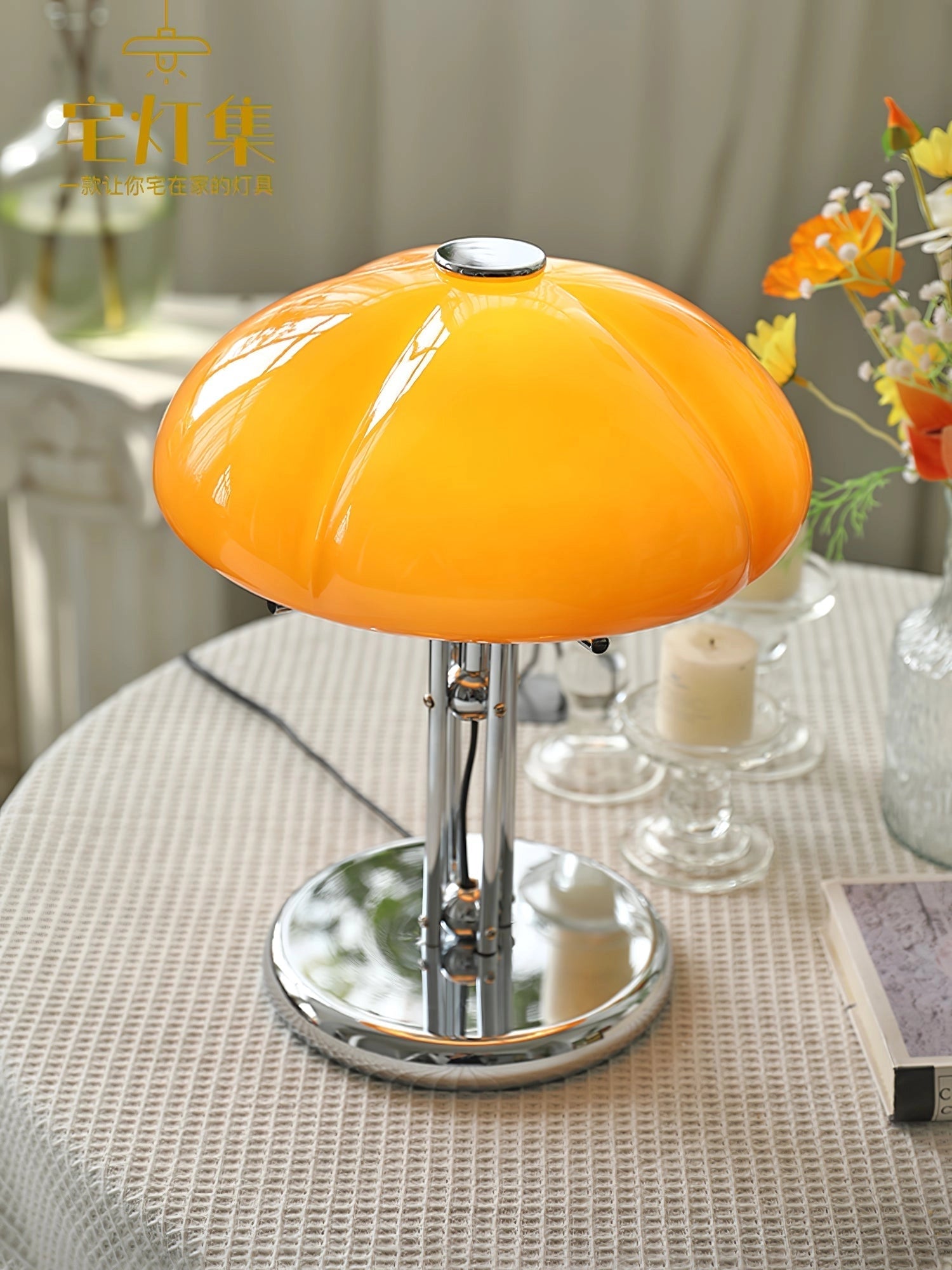 Modern orange globe table lamp with chrome base emitting warm light, placed on a table next to a plant and decorative tableware.