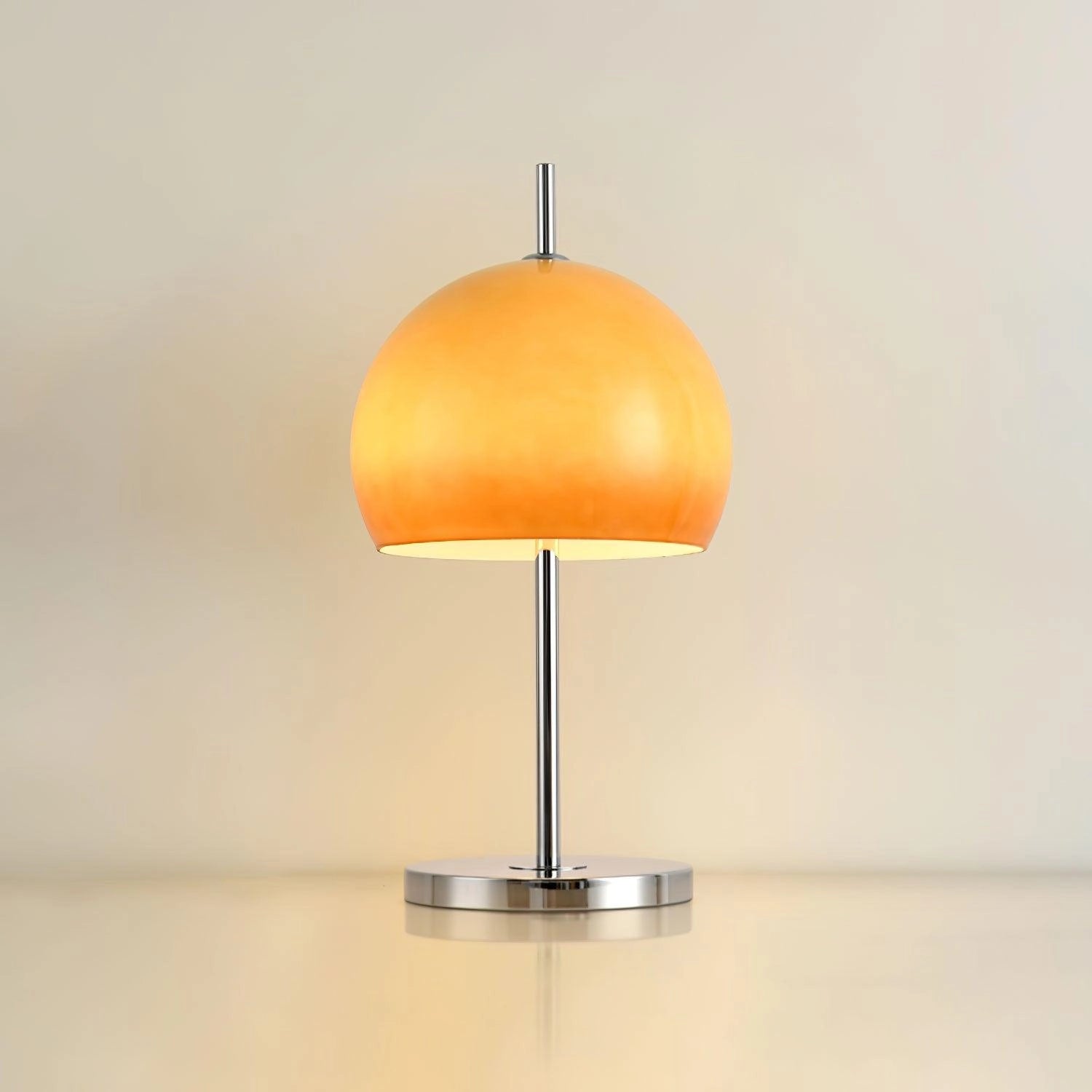 Modern orange globe table lamp with chrome base, emits warm light, placed on a wooden table next to a green plant.