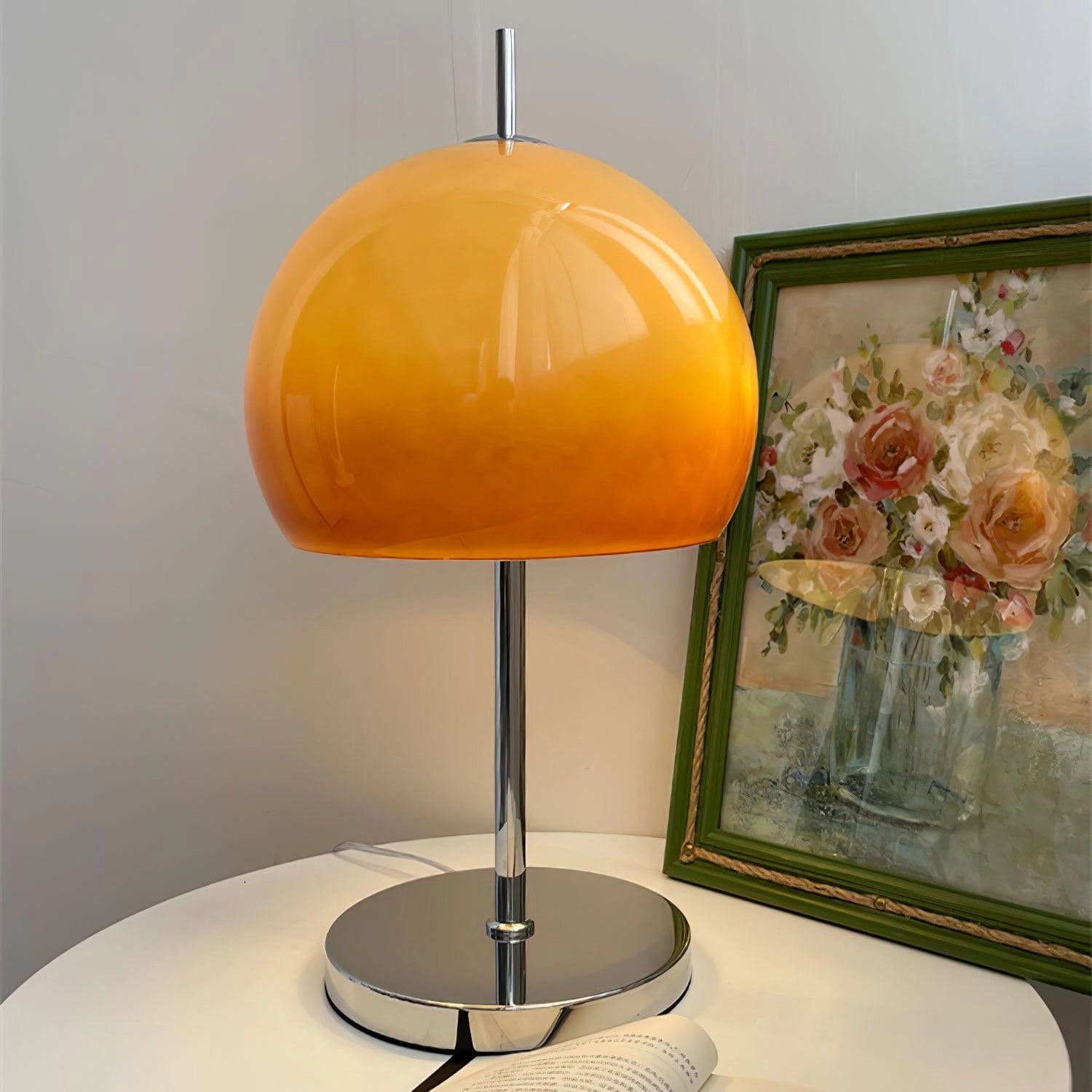 Modern orange globe table lamp with chrome base emitting warm light, placed on a table alongside a picture frame, plant, and decorative items.