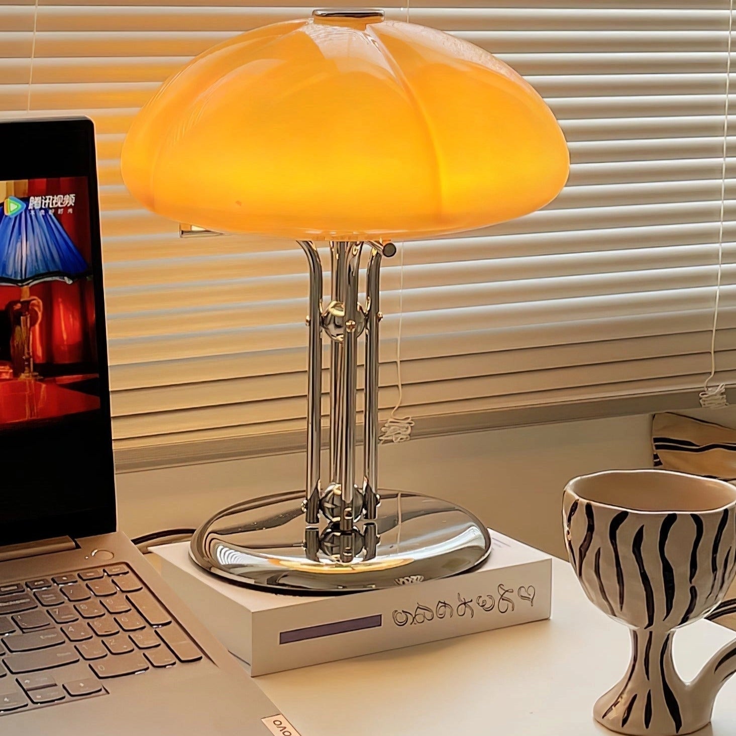 Modern orange globe table lamp with chrome base emitting warm light, placed on a wooden table next to a chair.