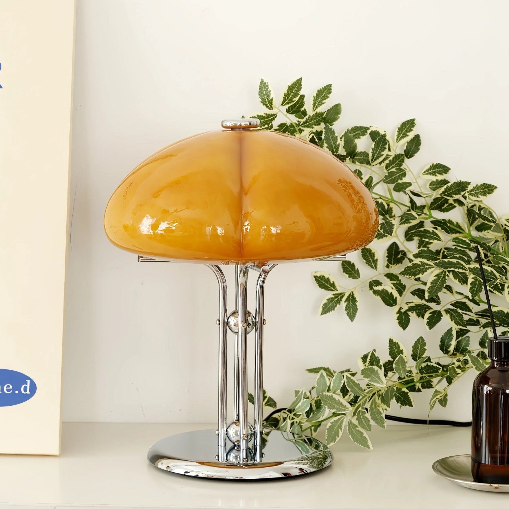 Modern orange globe table lamp with chrome base emitting warm light, placed on a table next to a small plant and glassware.