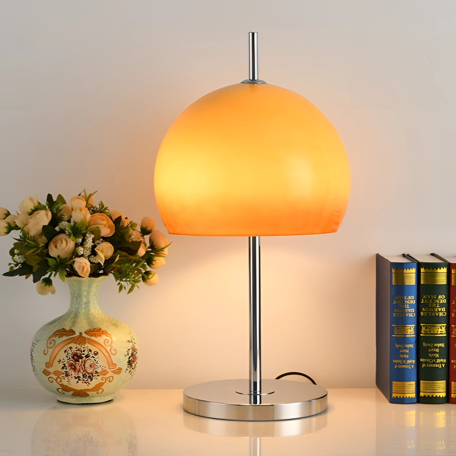 A modern orange globe table lamp with a chrome base emitting warm light, placed on a table near a vase with flowers and other decorative items.