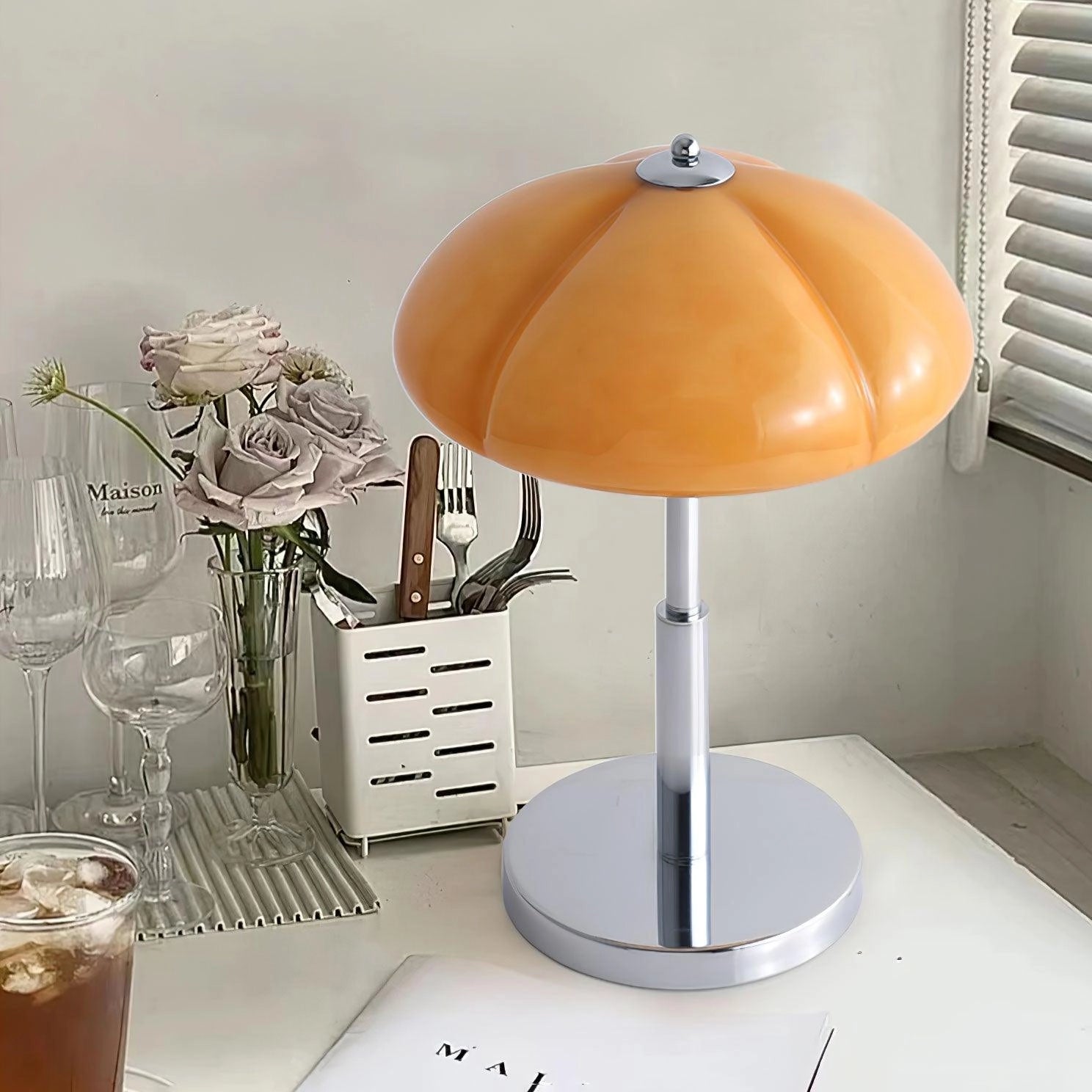 Modern orange globe table lamp with chrome base emitting warm light, placed on a wooden table with decorative elements including a plant and tableware in a stylish interior setting.