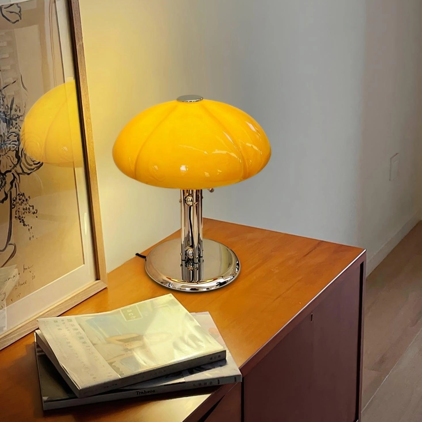 Modern orange globe table lamp with a chrome base, emitting warm light, placed on a wooden table next to a small potted plant in a stylish interior setting.