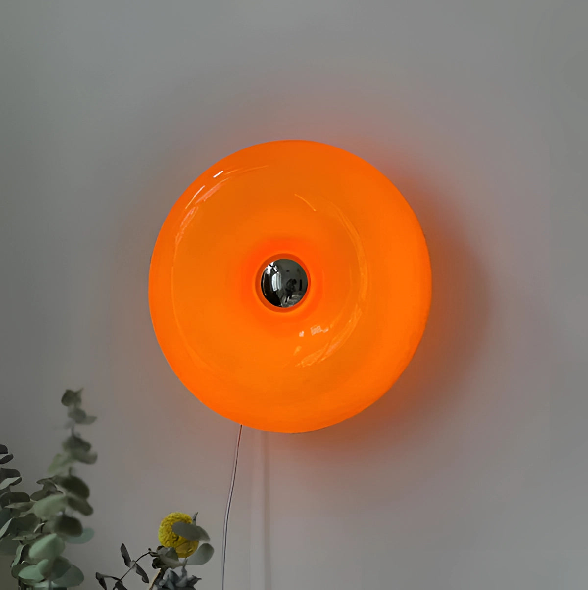Circular modern wall sconce with a vibrant orange shade, showcasing a contemporary design. The fixture is mounted on a wall with a small green plant nearby, adding a touch of natural decor to the scene. The circular shape and warm amber glow create an artistic and inviting ambiance.