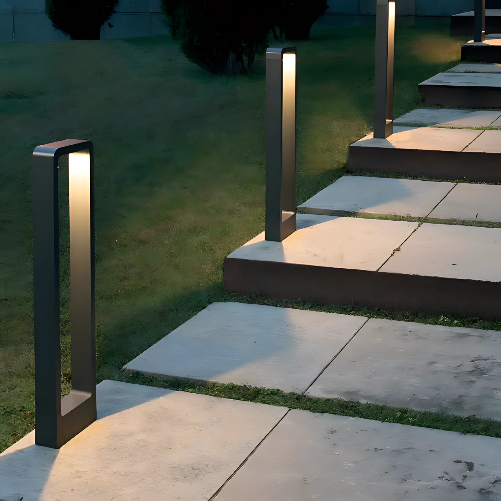 Modern outdoor LED pathway lights illuminating a garden path, surrounded by lush green grass and plants, with a clear view of the road surface. The lights have a sleek rectangular design and are made from weatherproof aluminum, seamlessly blending into the landscaped environment.