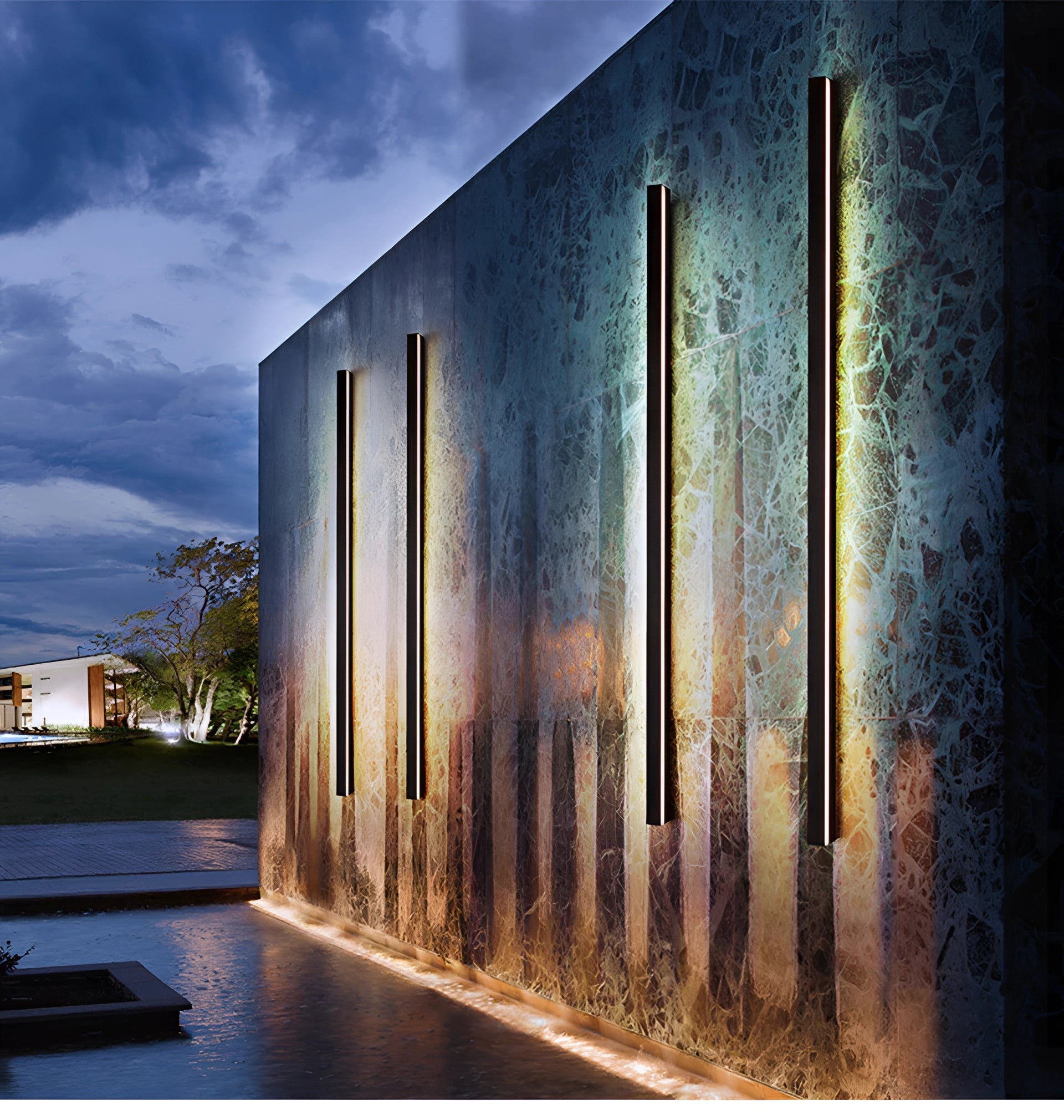 Modern outdoor LED wall light with a slim, minimalist design mounted on a building exterior, under a clear sky with some clouds, surrounded by elements of wood and plants.