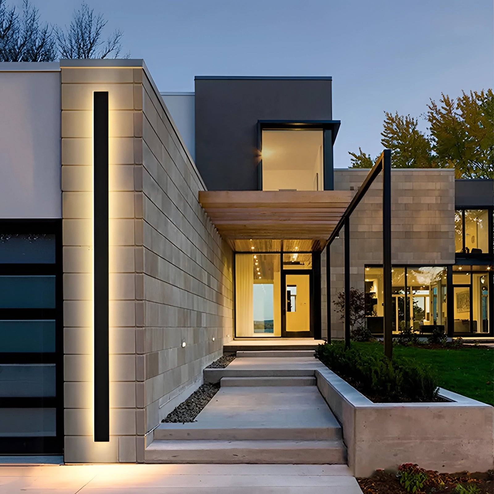 Modern outdoor LED wall light in a slim, minimalist design with a black finish, mounted on a building facade in a residential area. The light emits a neutral glow, complementing the surrounding architecture and nearby plants, under a clear sky.