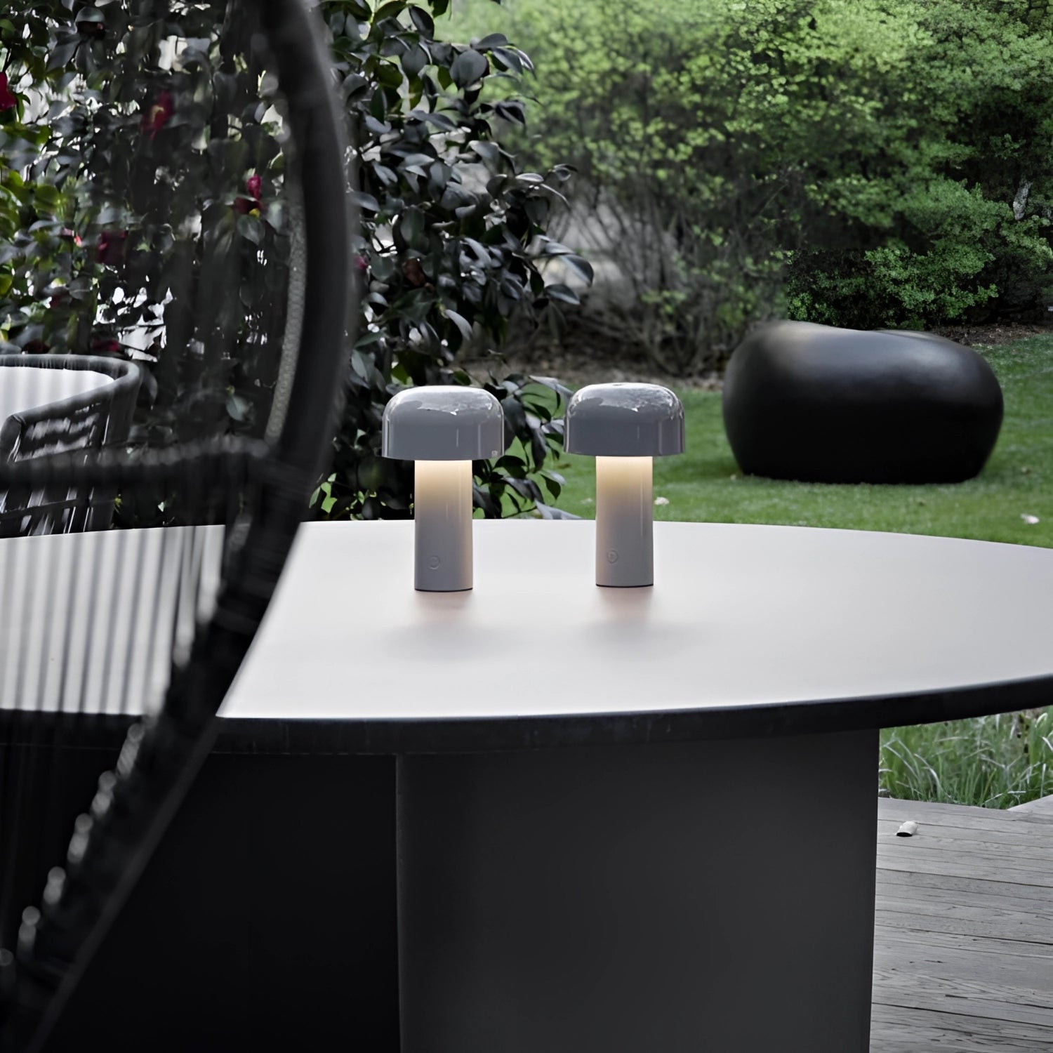 Modern portable wireless LED lamp with a sleek design and touch control, placed on a wooden outdoor table surrounded by greenery, with a potted plant nearby.
