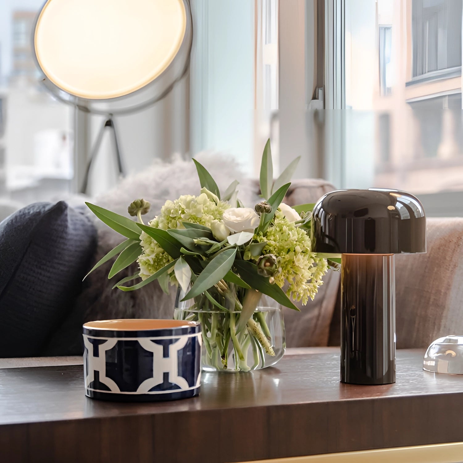 Modern portable wireless LED lamp with a sleek design, placed on a table near a flower and drinkware, with natural light coming through a window in the background.