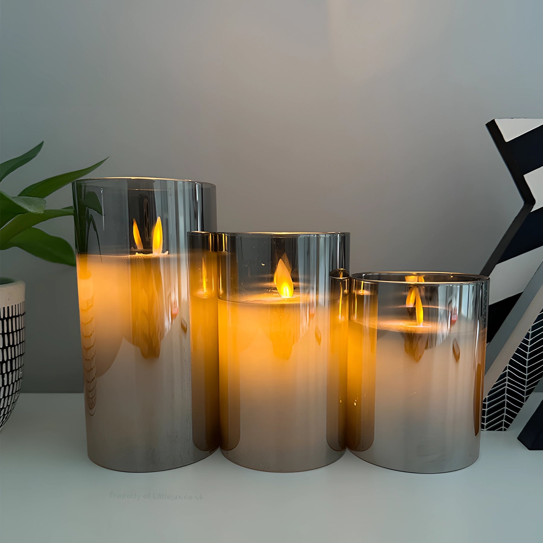 Elegant minimalist decor featuring modern reflective glass candle holders surrounded by green plants and set against a wooden surface.