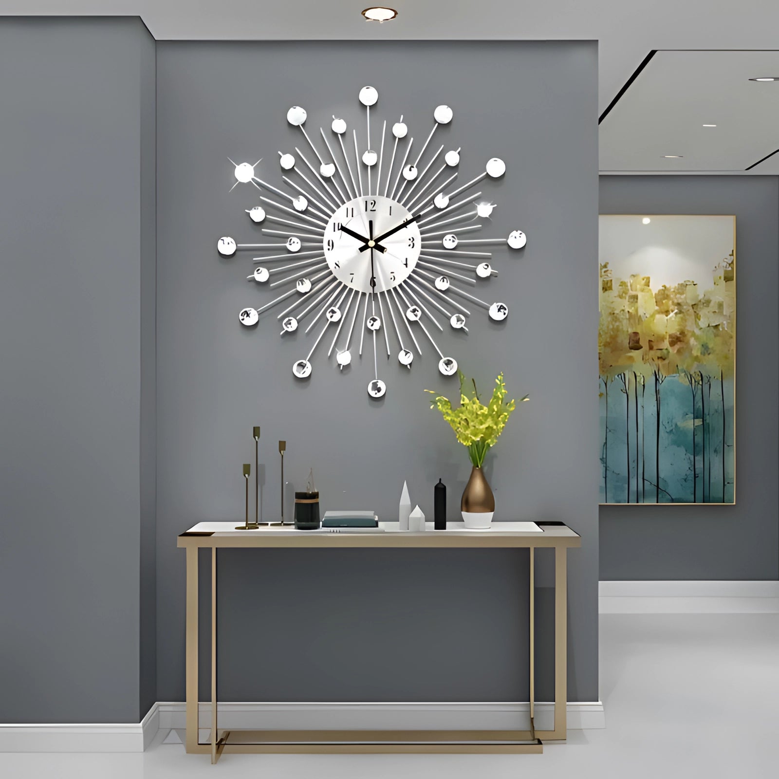 Modern starburst silver wall clock with a mid-century design, mounted on a grey wall. The clock features a unique starburst pattern with sleek silver spokes. Below, a wooden table holds a vase with branches, adding a touch of natural decor to the interior design.
