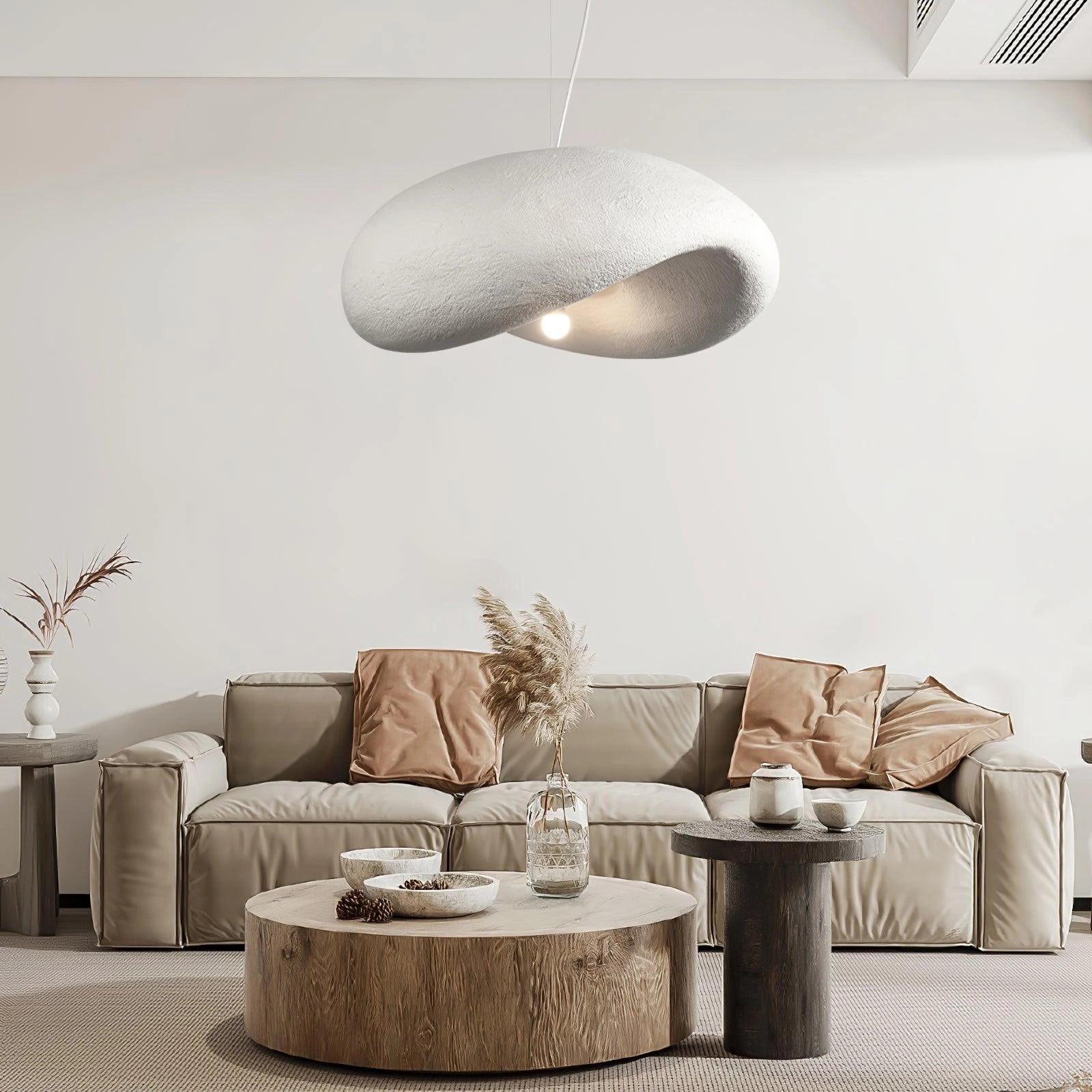 Modern textured pendant light with a minimalist pebble design and stone finish, hanging in a stylish interior setting with a brown couch, beige and grey elements, and a wooden floor.