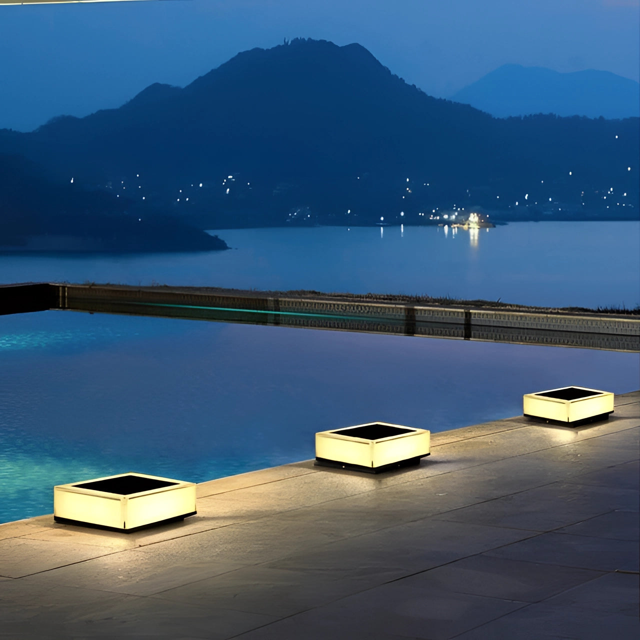 Modern waterproof LED cube seats illuminated at night near a tranquil body of water with a mountainous backdrop under a clear sky.