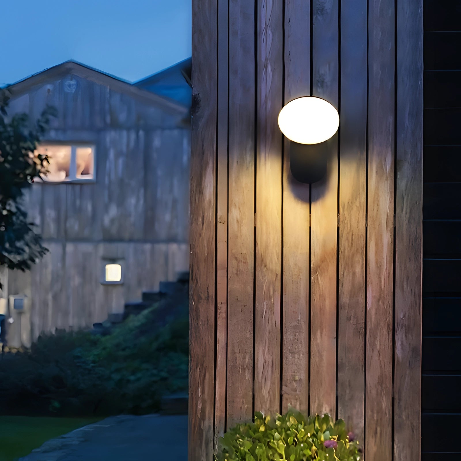 Modern weather-resistant LED wall lantern with motion sensor mounted on the exterior of a building, surrounded by plants and flowers, with a view of the sky and additional architectural elements like windows and siding visible.