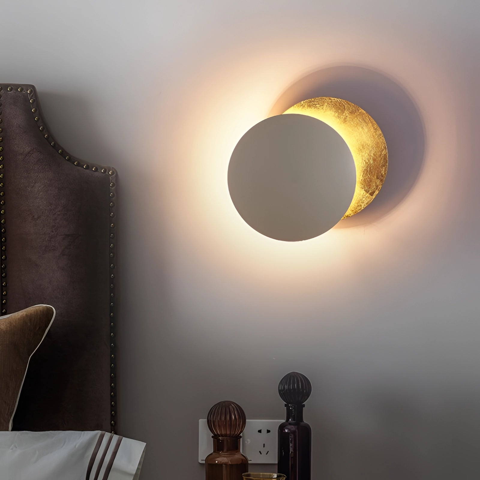 Luxury gold moon-phase wall light, designer lunar sconce with circular shape, resembling a moon, casting soft tints and shades; mounted on a wooden backdrop with artistic and astronomical elements, providing comfort and enhancing room aesthetics.