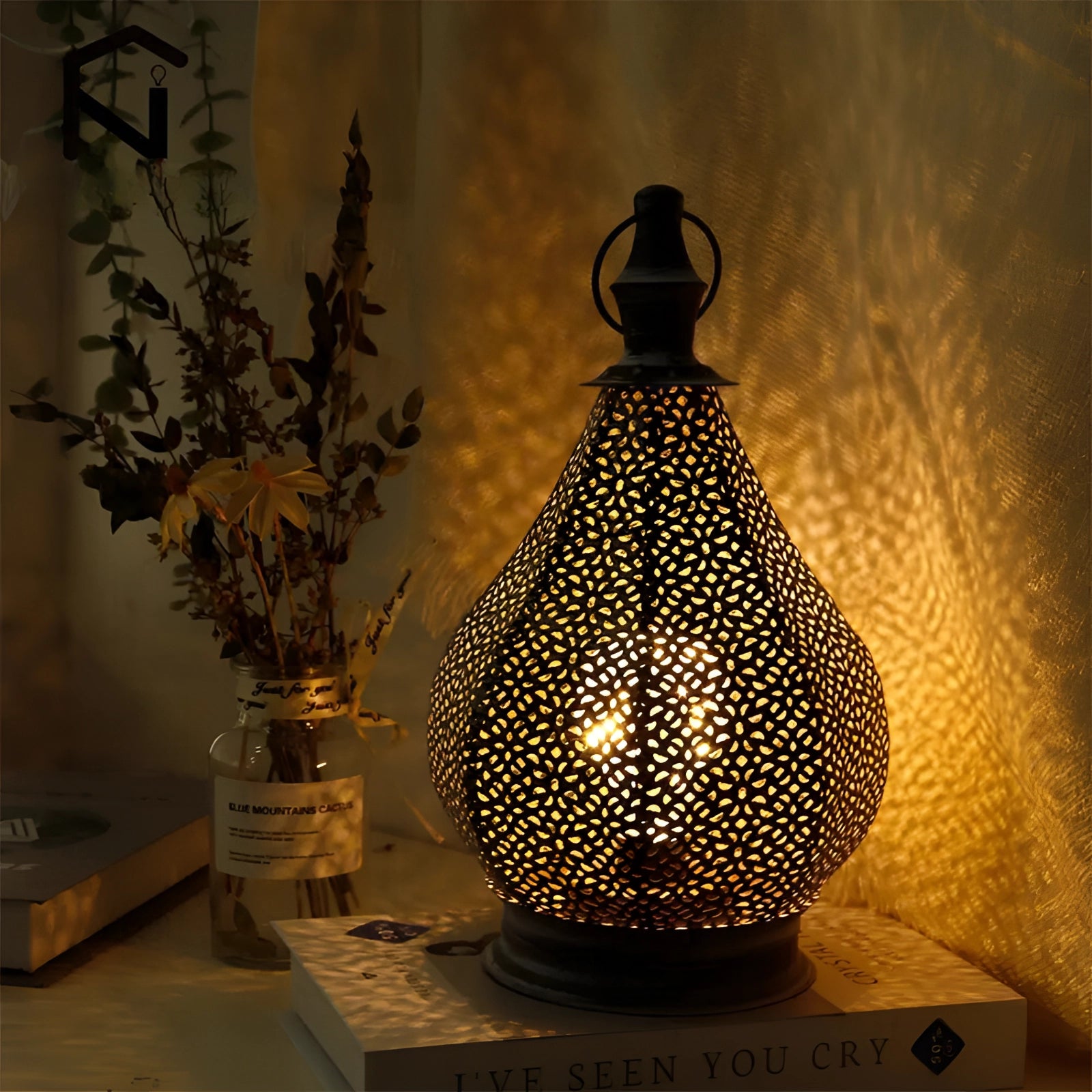 Moroccan metal table lamp with intricate patterns, casting ambient lighting in an interior setting. The lamp is placed on a wooden surface, surrounded by a window and a plant, creating an ethnic and cozy atmosphere.
