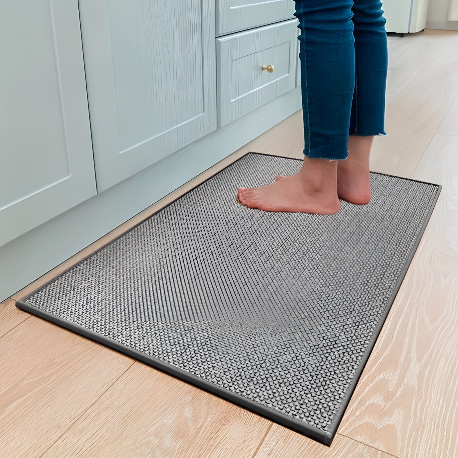 Non-slip gray kitchen mat with a durable, washable sisal surface, measuring 50 x 80 cm, placed on a hardwood floor beneath wooden cabinetry. The rectangular rug adds a touch of style to the kitchen decor while providing comfort and safety.