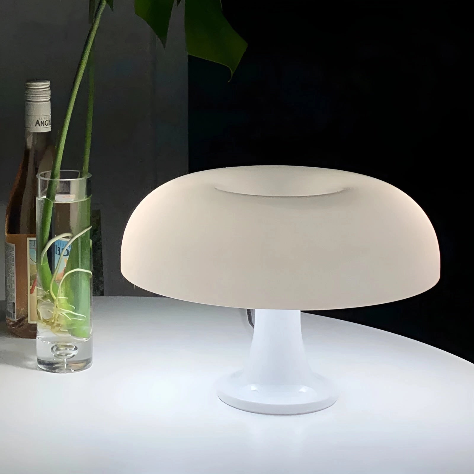 Orange mushroom lamp with a retro 70s style design, featuring a white base, placed on a table next to a small plant and a bottle, showcasing a mid-century lighting aesthetic.