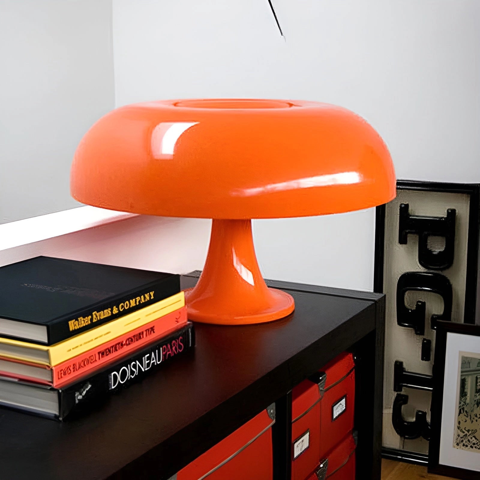 Orange mushroom lamp in retro 70s style with mid-century design, placed on a wooden table or desk, surrounded by shelves and cabinetry in a home or office setting.