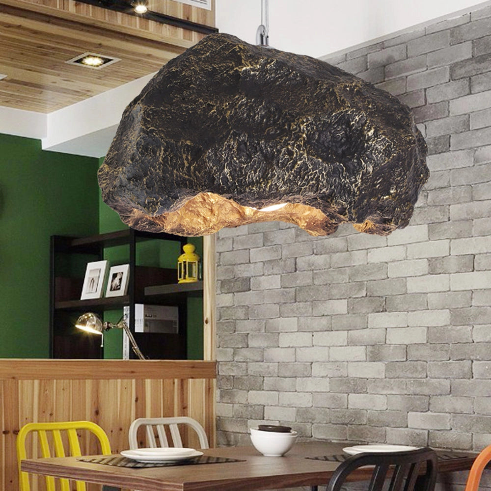 Organic stone pendant light hanging from ceiling in a modern industrial interior setting, surrounded by wooden furniture including a table and chairs, with artistic elements and wood flooring.