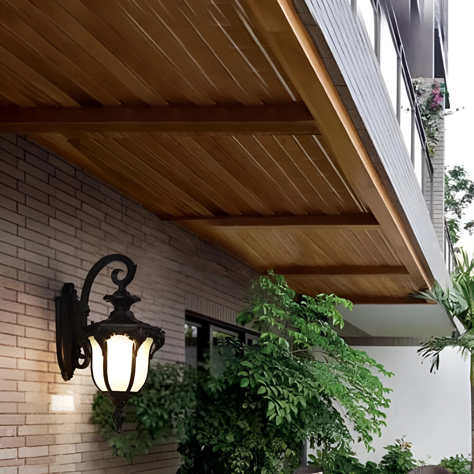 Outdoor wall lantern with vintage bronze finish mounted on a wooden exterior wall, emitting warm light near a window, surrounded by plants and part of a house structure.