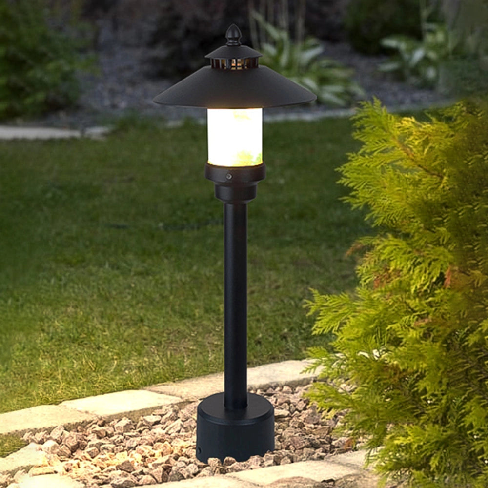 Pagoda-inspired weather-resistant outdoor path light positioned among grass and plants, featuring a metal design with a lamp emitting a warm glow, suitable for garden or pathway illumination.