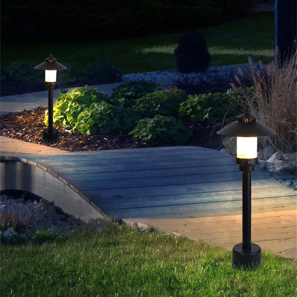 Pagoda-inspired black outdoor path light standing on an asphalt sidewalk surrounded by green grass and plants, illuminated by sunlight.