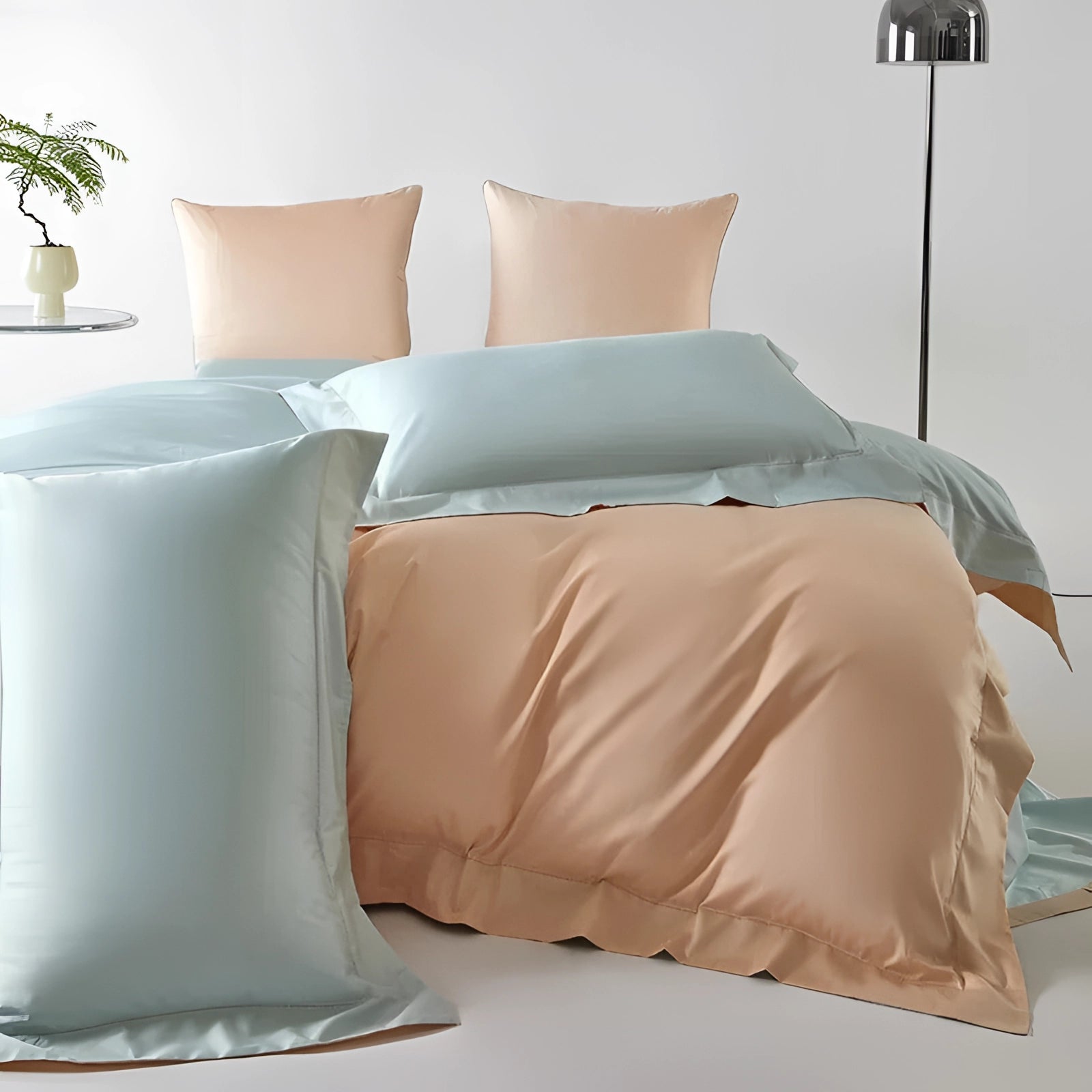 Pastel reversible satin duvet set in peach and aqua displayed on a bed, complemented by pillows, a flowerpot, a wooden floor, and soft lighting, creating a comfortable and stylish bedroom interior design.