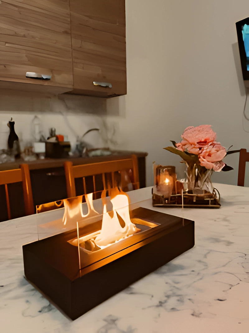 Portable bio-ethanol tabletop fireplace with a sleek design, set on a wooden table in a modern living room with hardwood flooring. The fireplace emits a warm orange flame, complementing the contemporary interior decor.